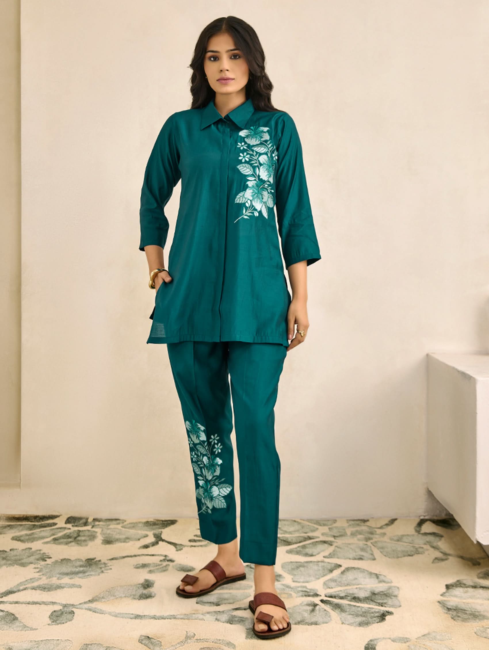 Woman wearing a teal outfit with floral embroidery on a neutral background