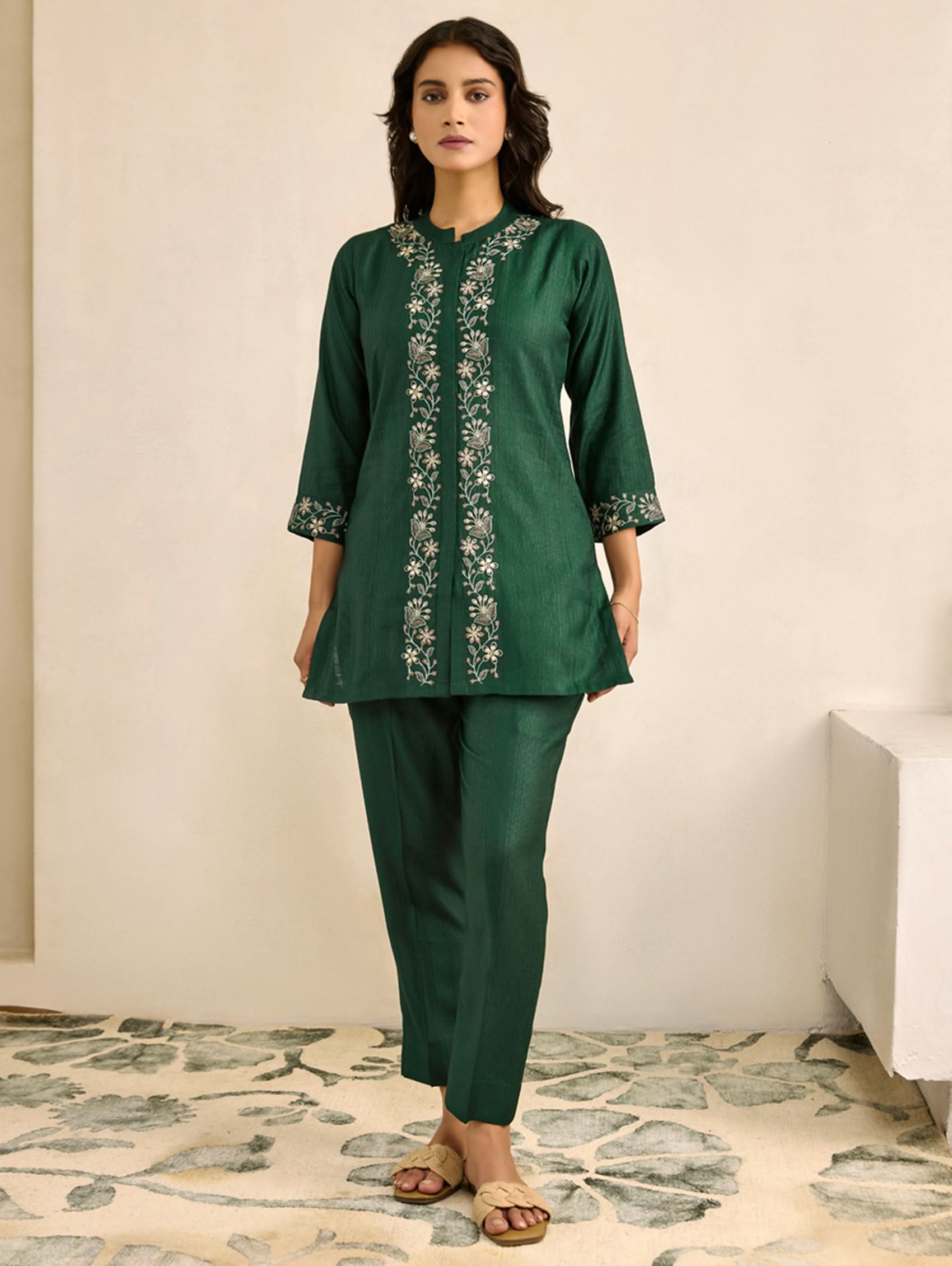 Woman wearing a green embroidered outfit standing in a room with a floral rug.