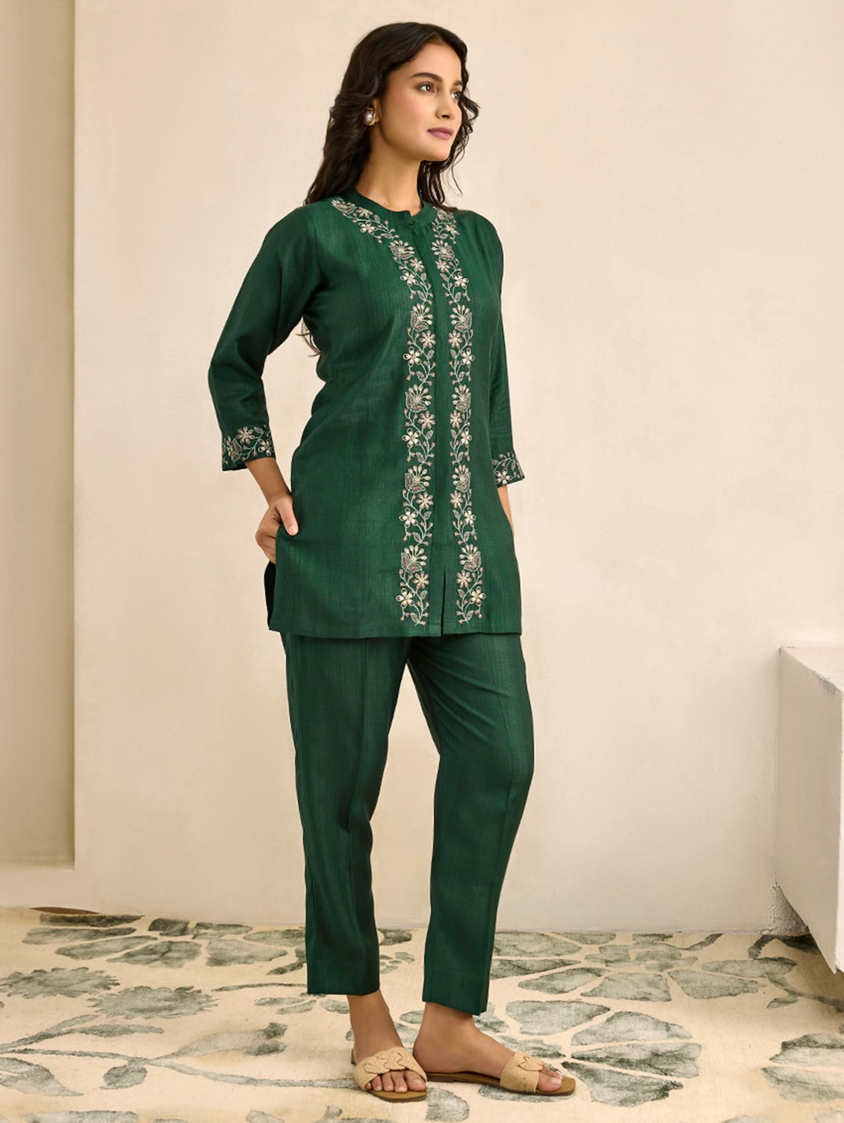 Woman wearing a green embroidered outfit standing in a room with a neutral background.