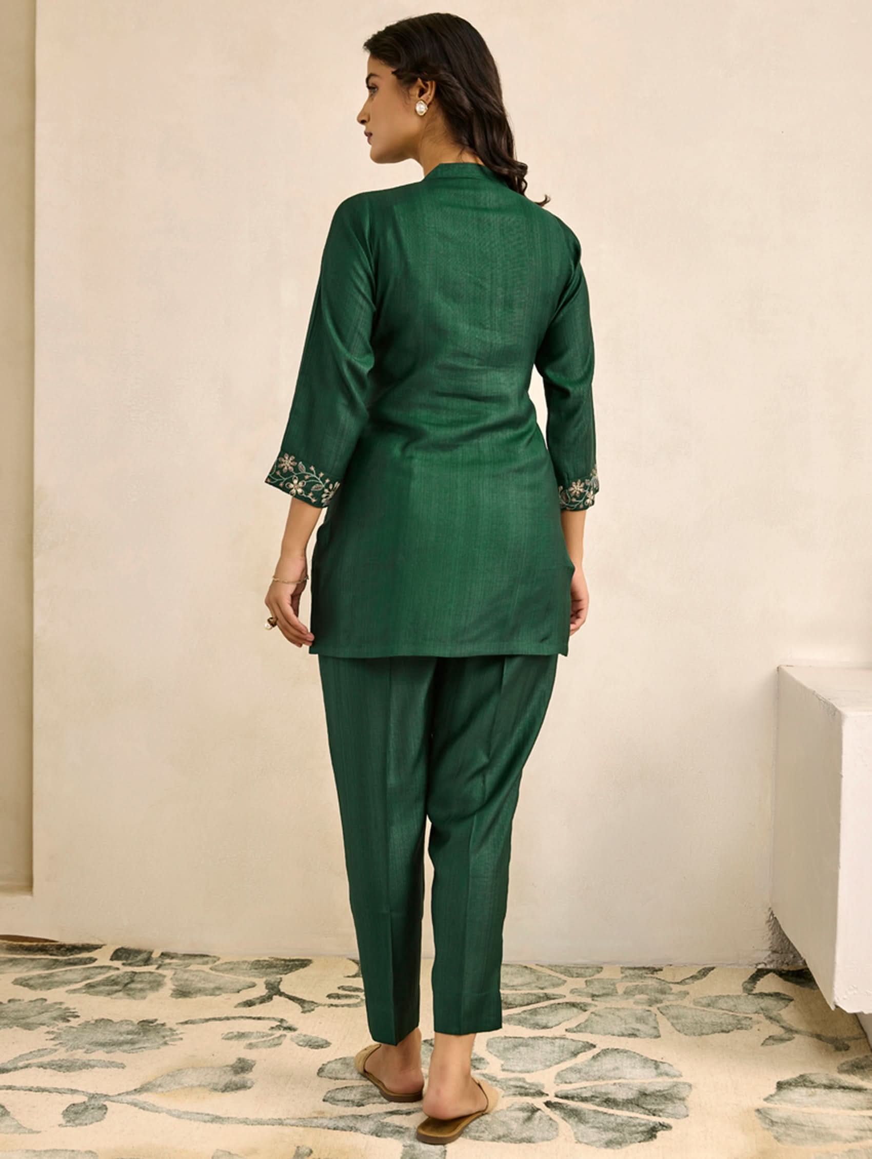 Woman wearing a green traditional outfit standing in a room with a light-colored wall and patterned rug.
