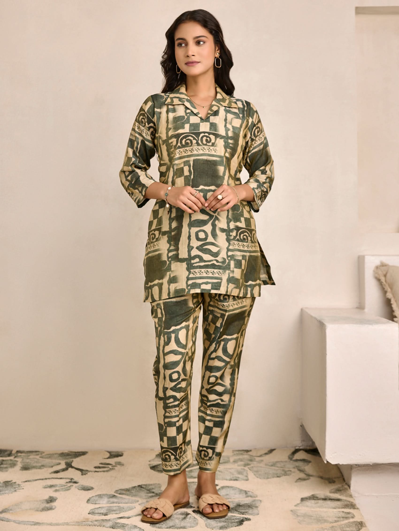 Woman wearing a green patterned outfit in a room with a white wall and a piece of furniture.