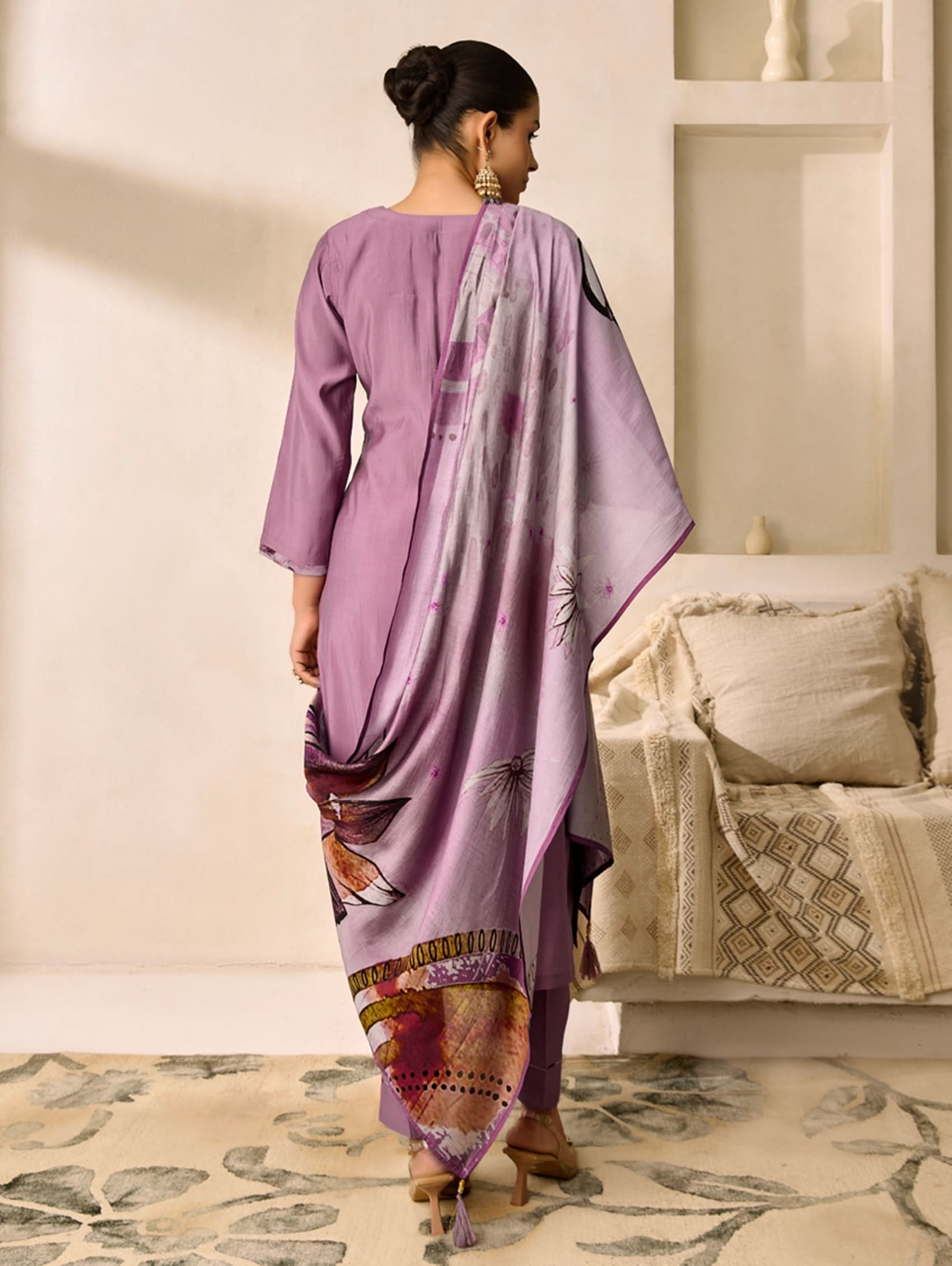Woman in a lavender traditional outfit with a patterned drape, standing in a room with a sofa and decorative pillows.
