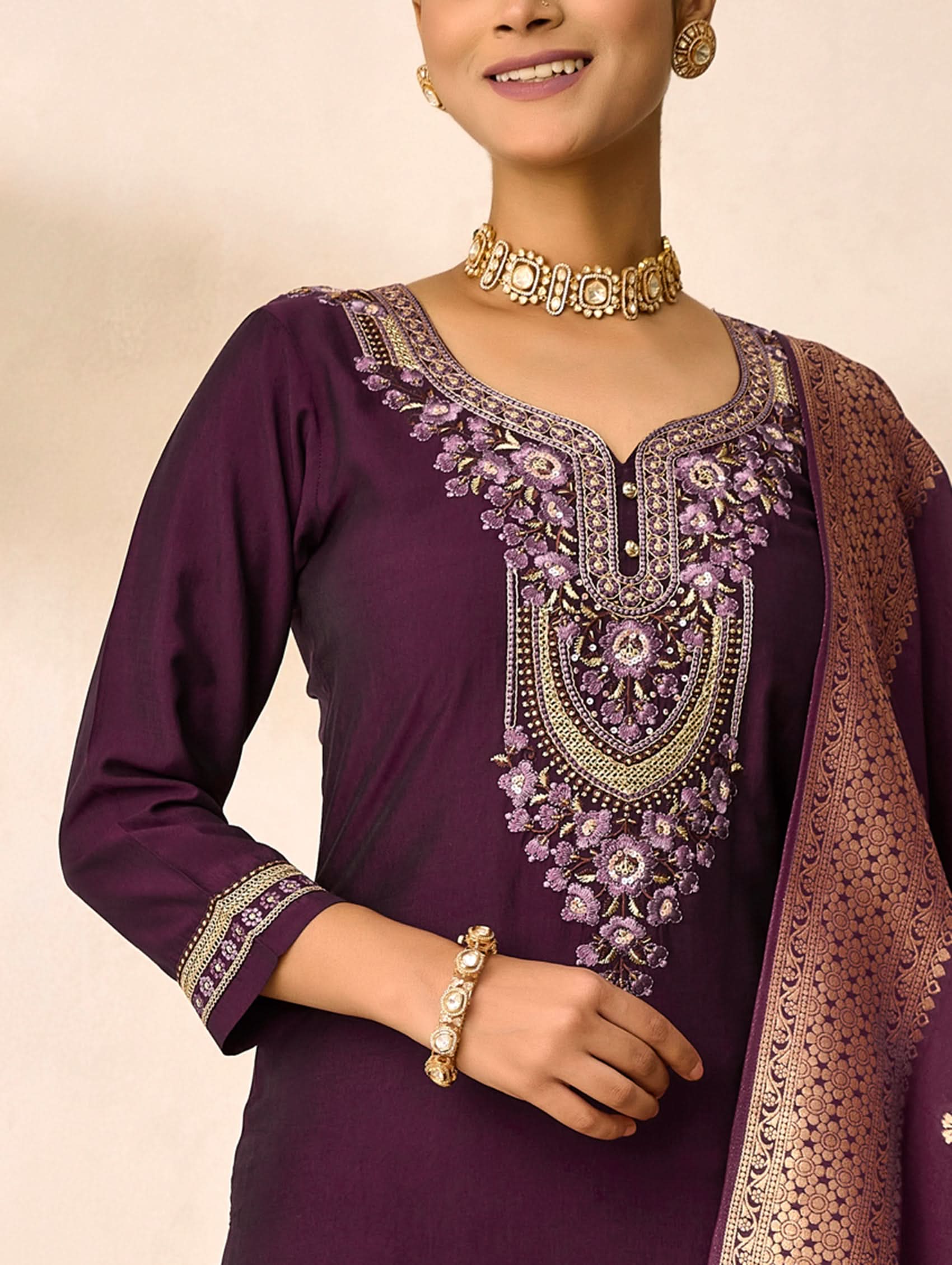 Woman wearing a purple embroidered traditional outfit with gold jewelry on a beige background