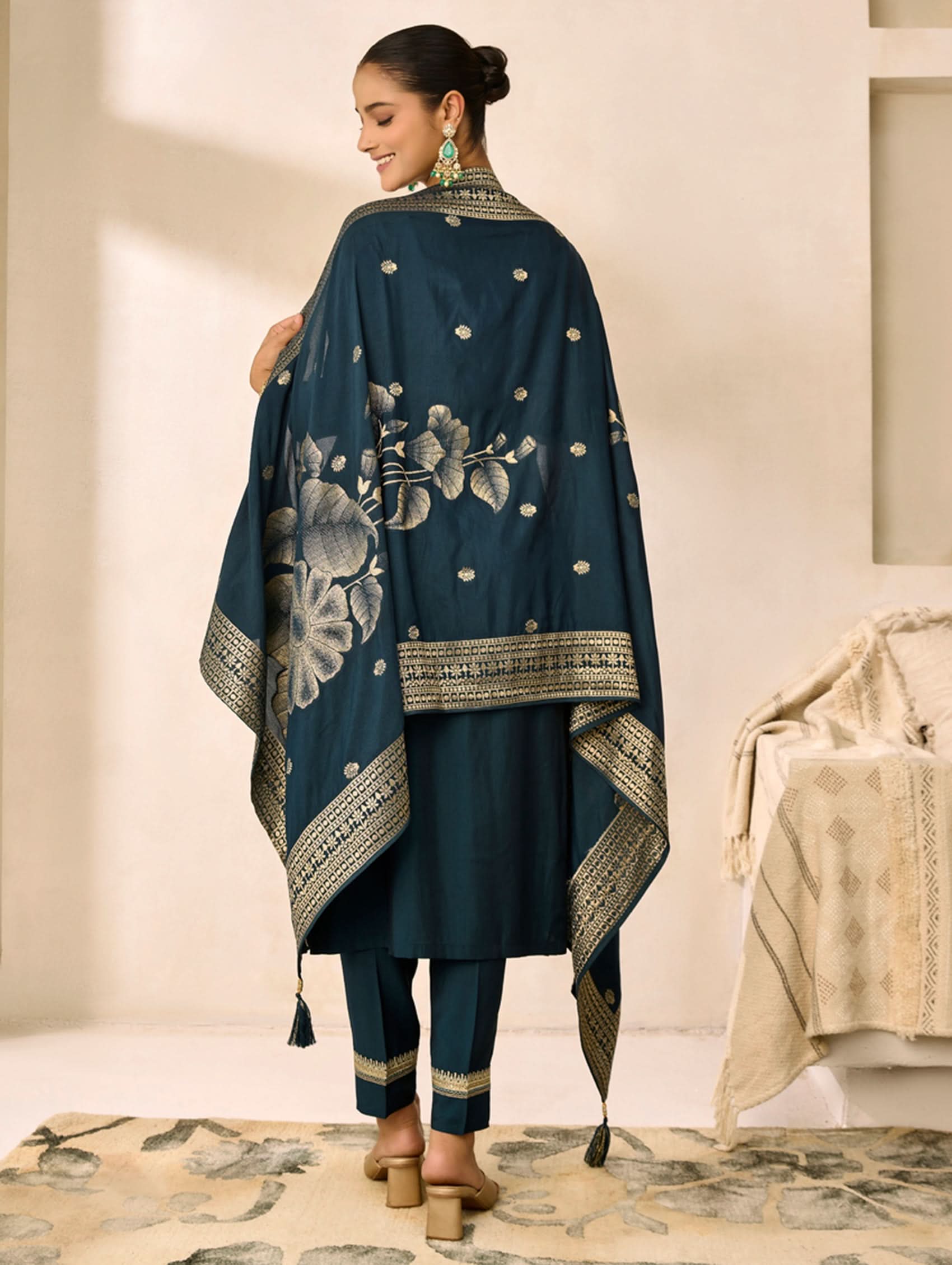 Woman wearing a teal embroidered outfit in a room with beige walls and a patterned rug.