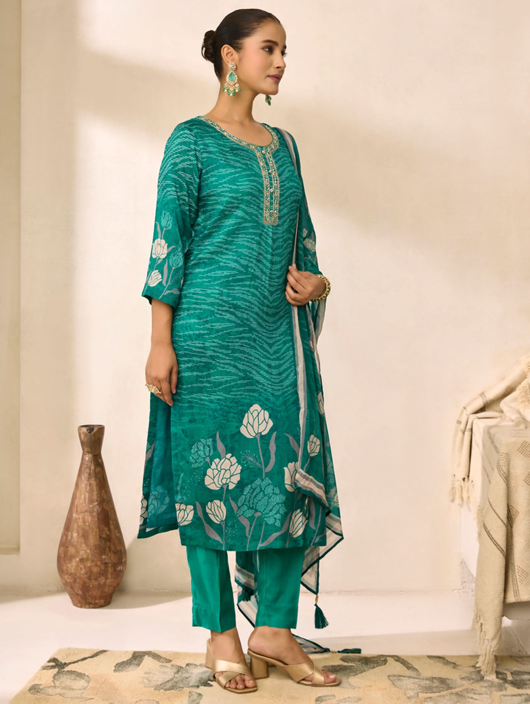 Woman wearing a teal embroidered outfit in a room with a vase and table.