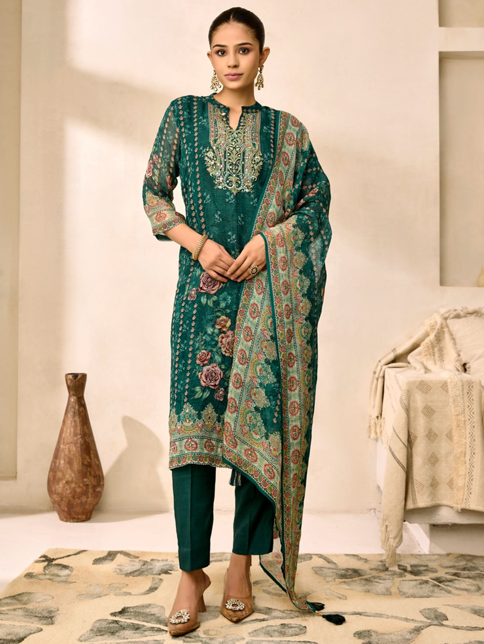 Woman wearing a green embroidered traditional outfit in a room with a vase and chair.