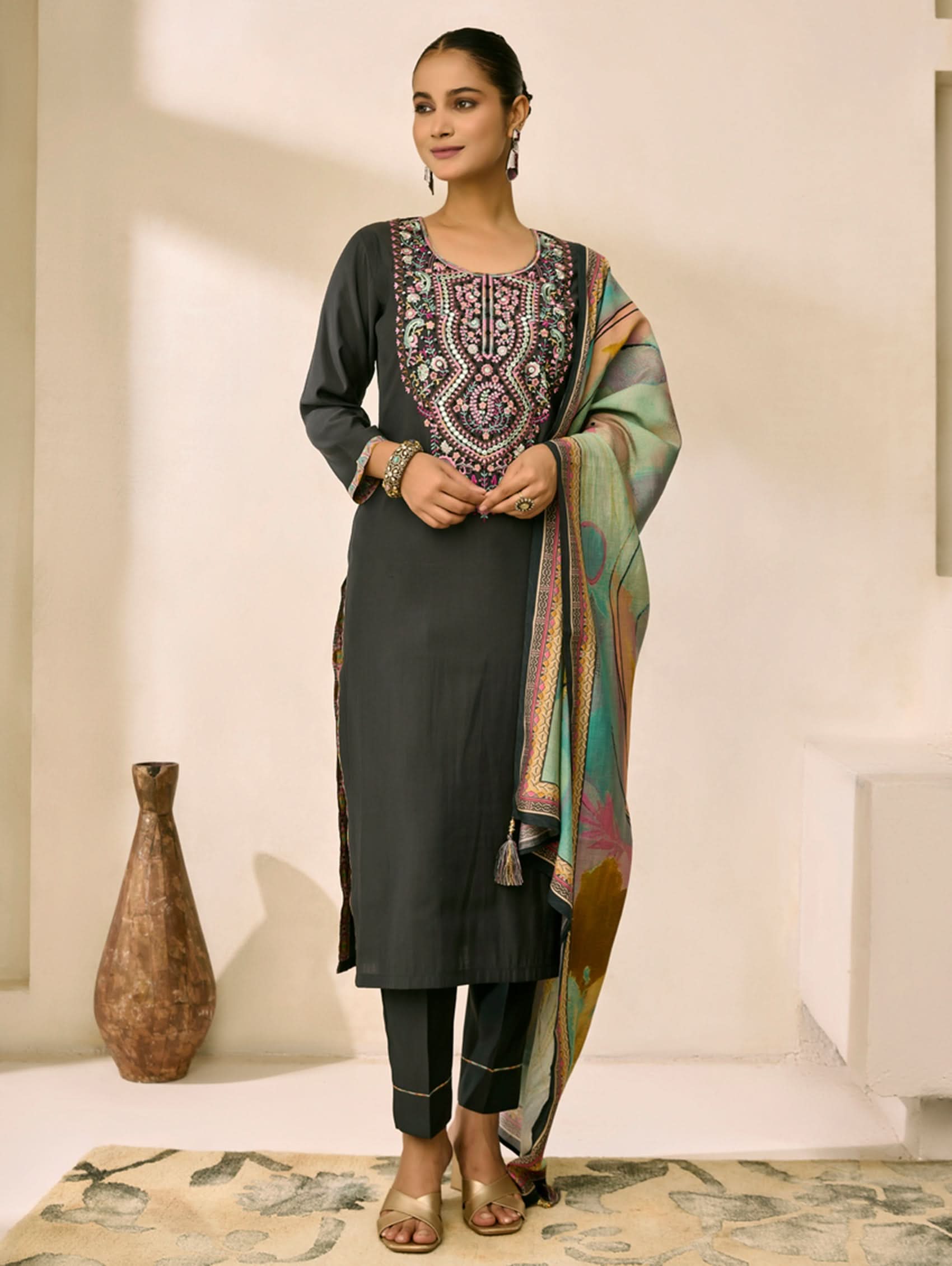 Woman wearing a black kurta with intricate embroidery and a colorful dupatta in a minimal indoor setting.