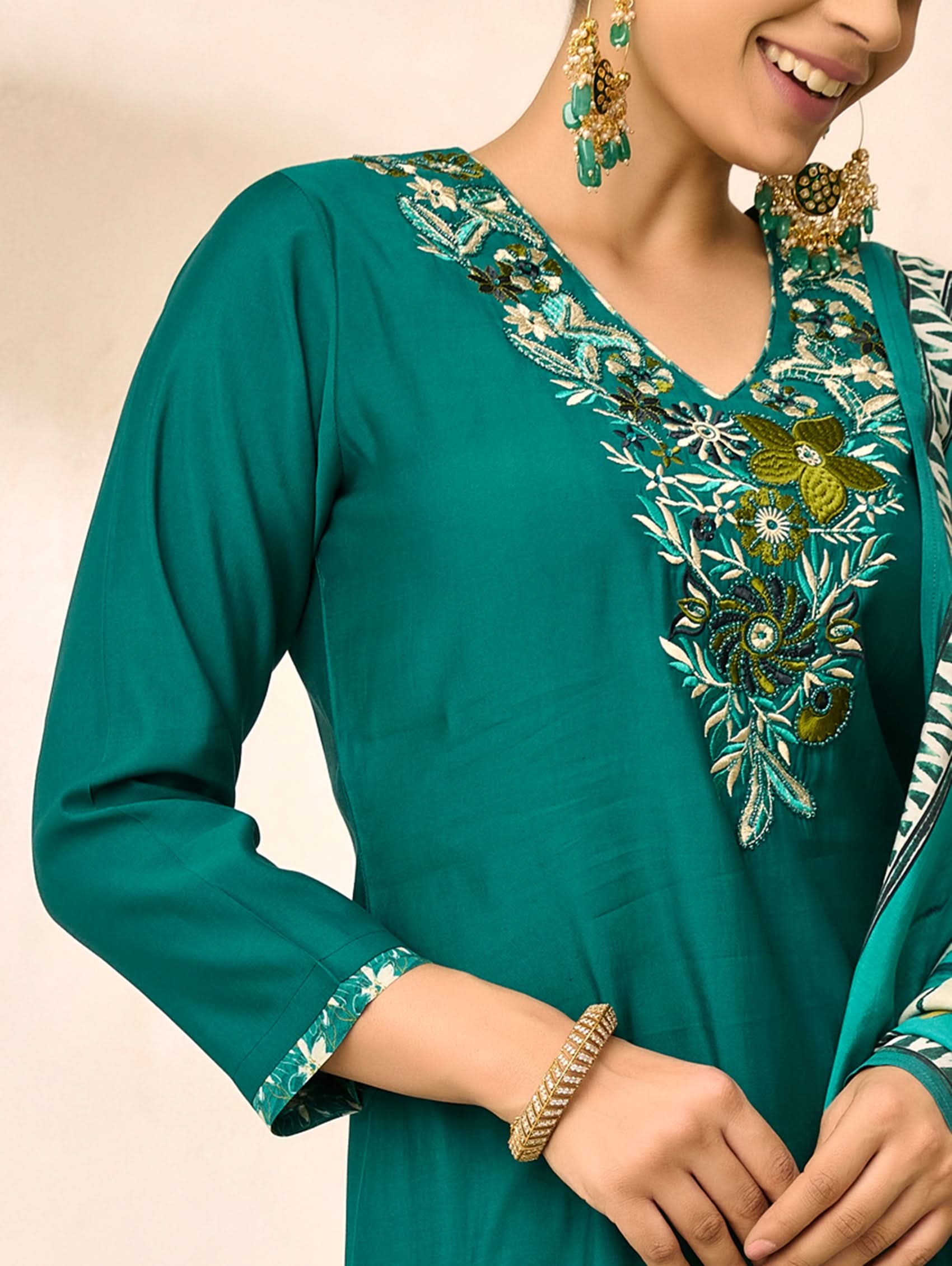 Teal embroidered traditional outfit with gold jewelry on a beige background