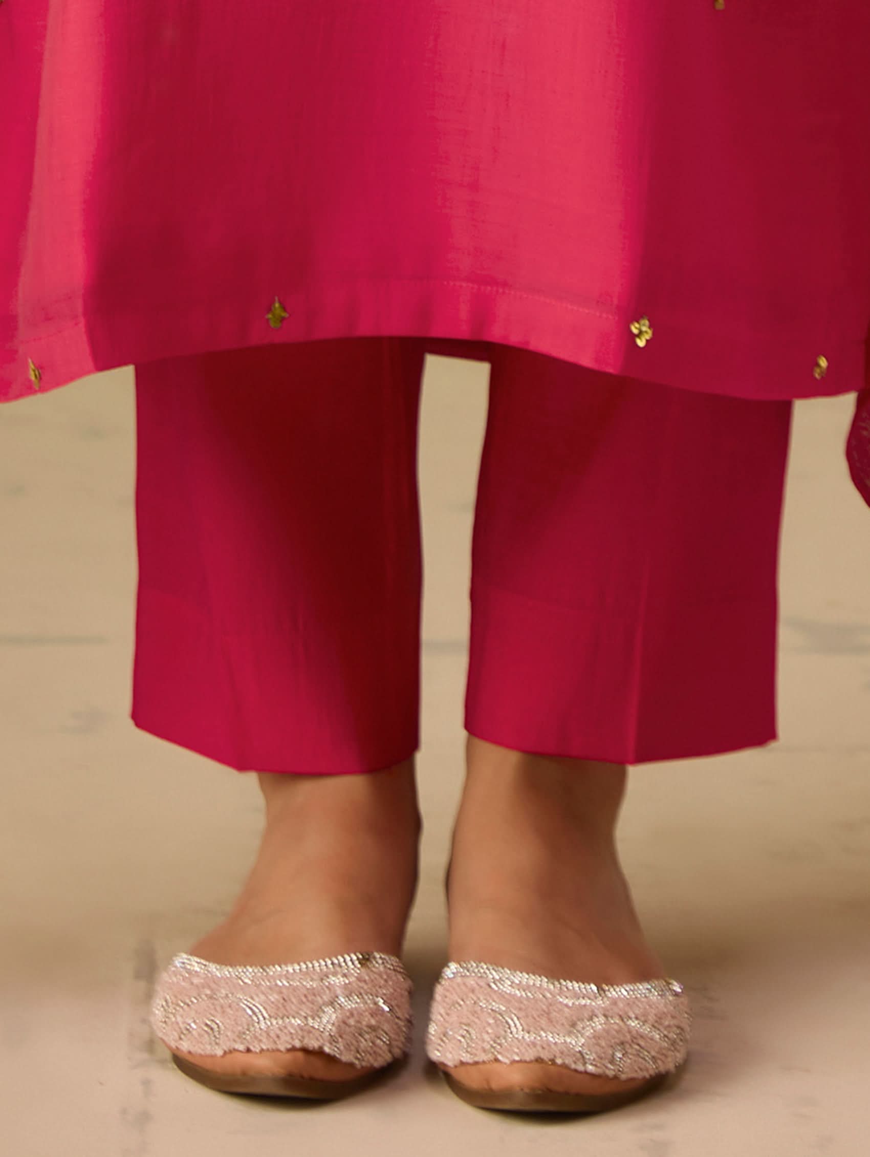 Person wearing a bright pink traditional outfit with matching pants and silver sandals on a neutral background