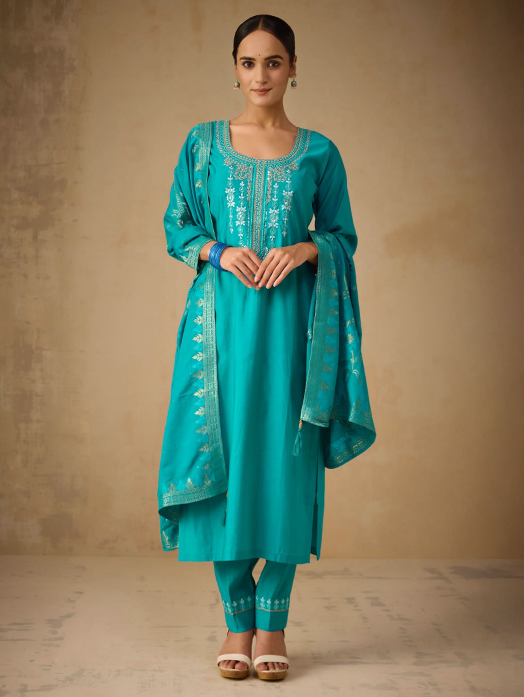 Woman wearing a teal embroidered outfit against a beige background