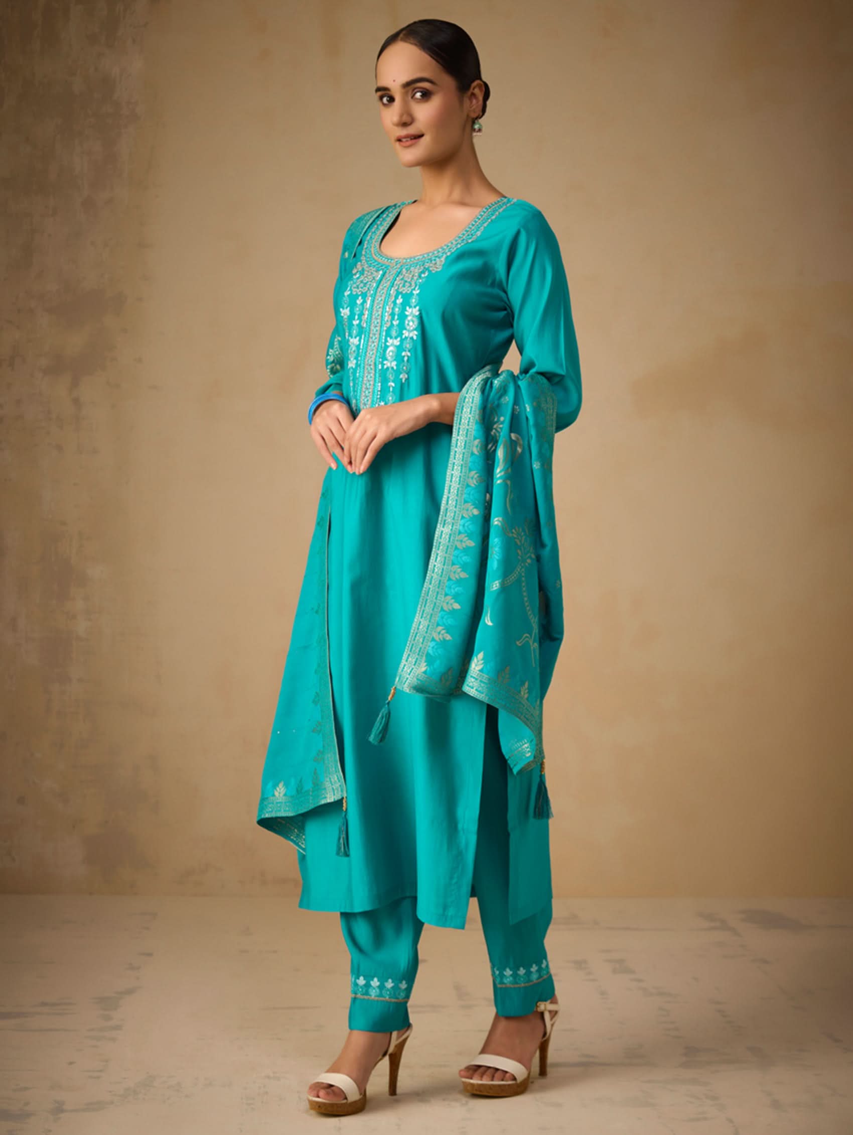 Woman wearing a turquoise traditional outfit with a matching dupatta against a beige background