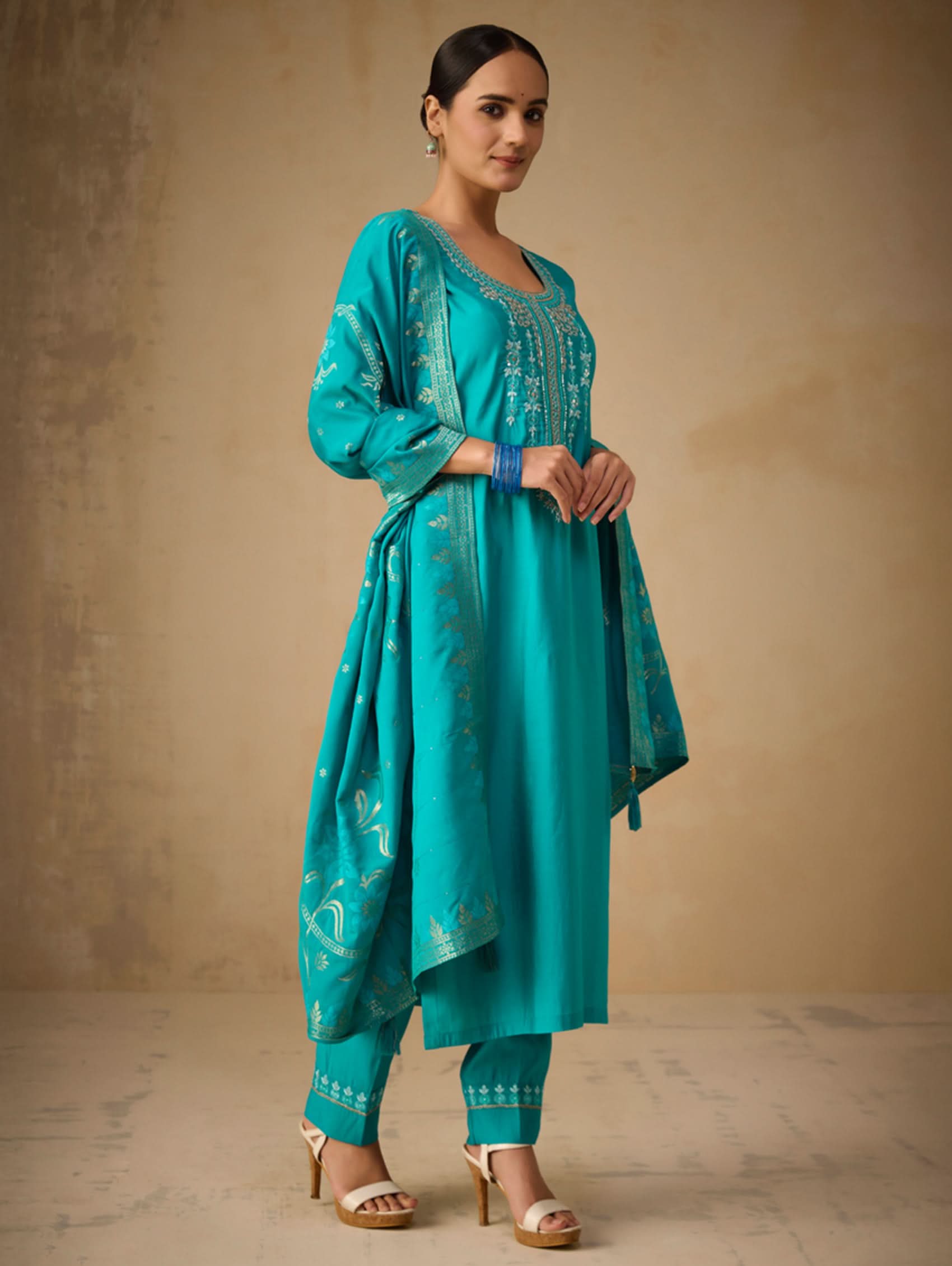 Woman wearing a teal traditional outfit with embroidery against a beige background