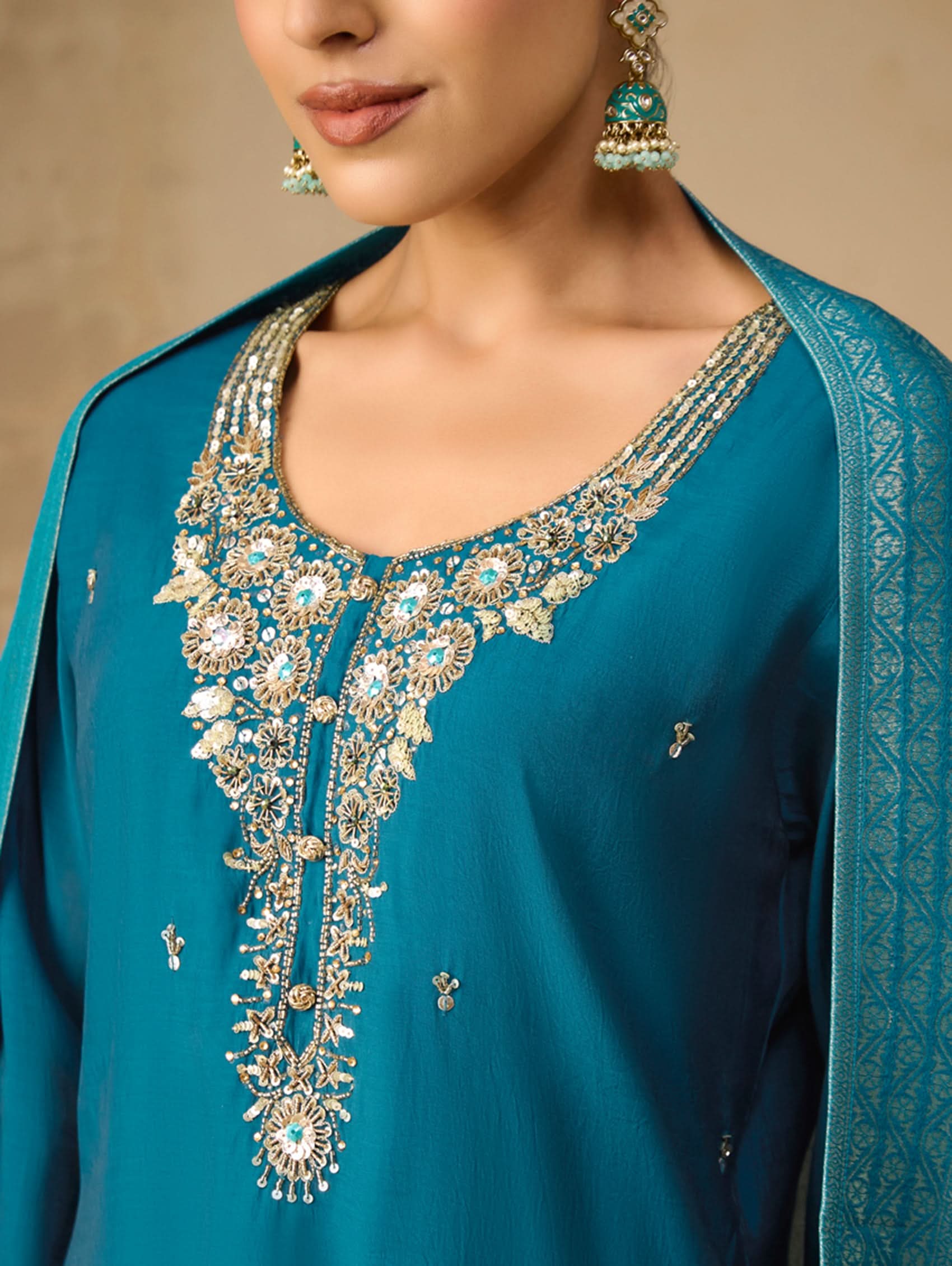 Close-up of a teal embroidered garment with gold detailing on a beige background
