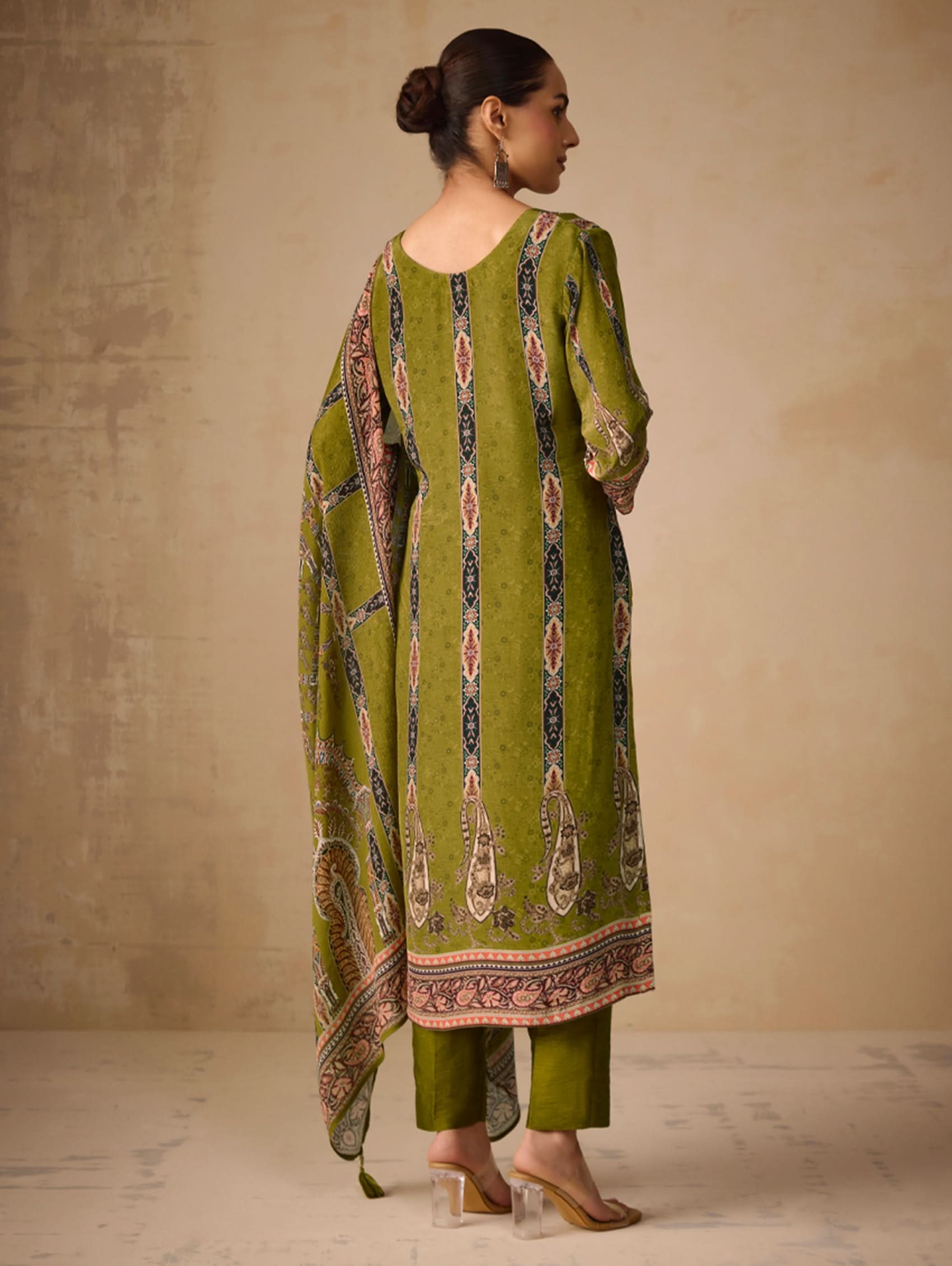 Woman wearing a green traditional outfit with a patterned shawl against a beige wall.