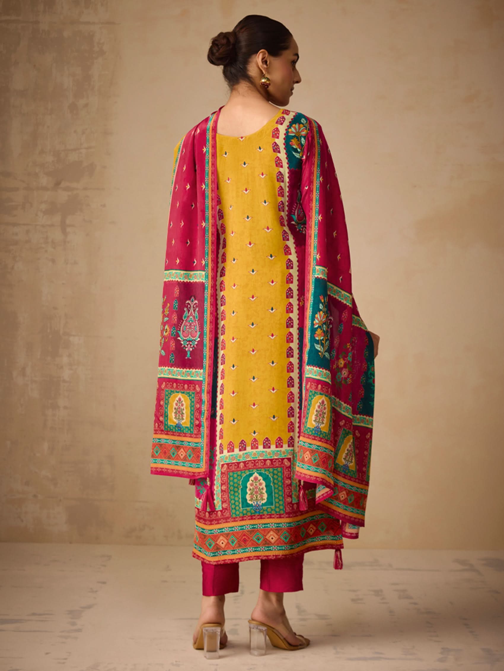 Woman wearing a colorful traditional outfit with a pink and multicolor dupatta against a beige wall.