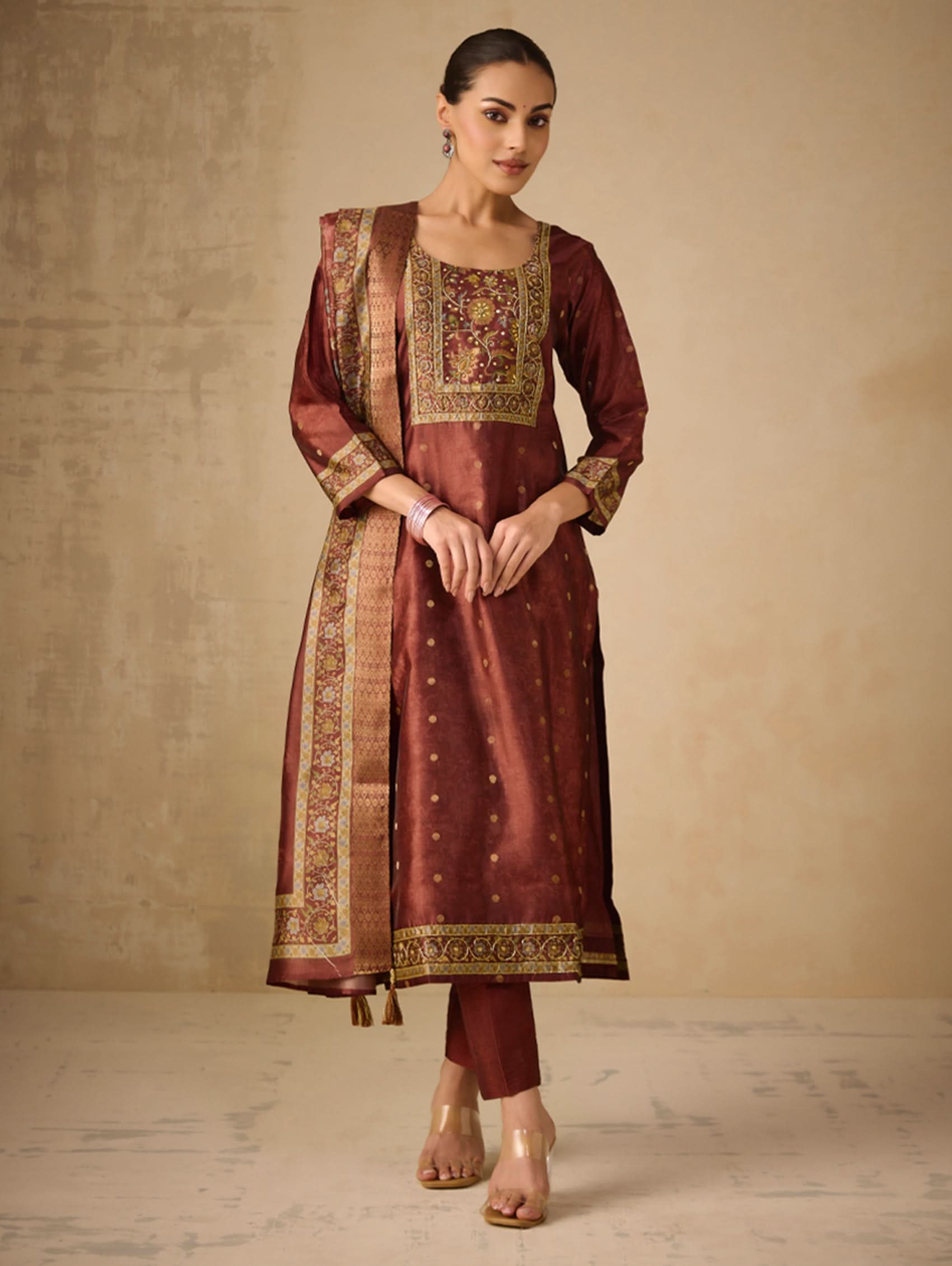 Woman wearing a traditional maroon outfit with gold embroidery against a beige background