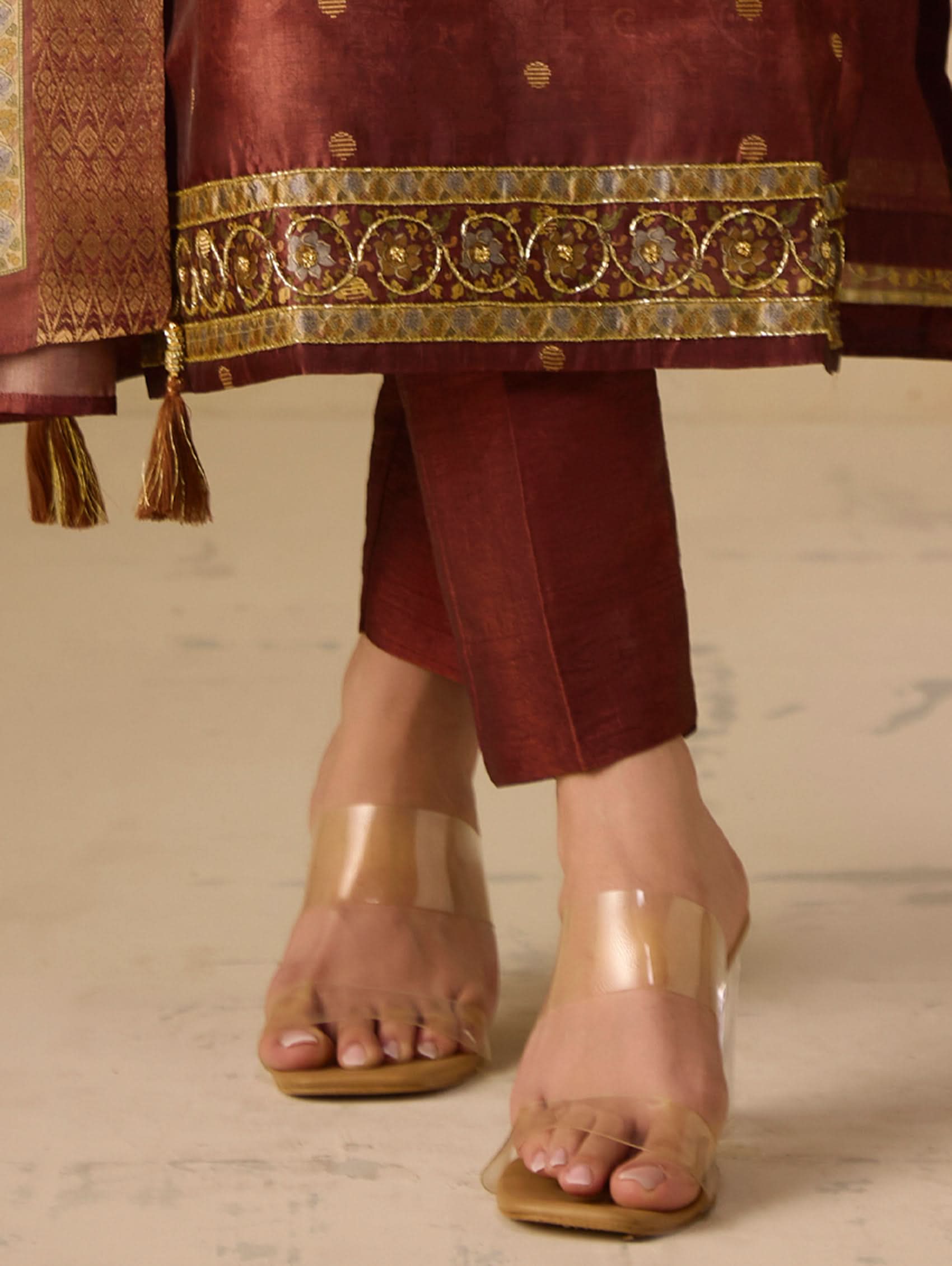 Maroon traditional outfit with gold embroidery and transparent sandals on a neutral background