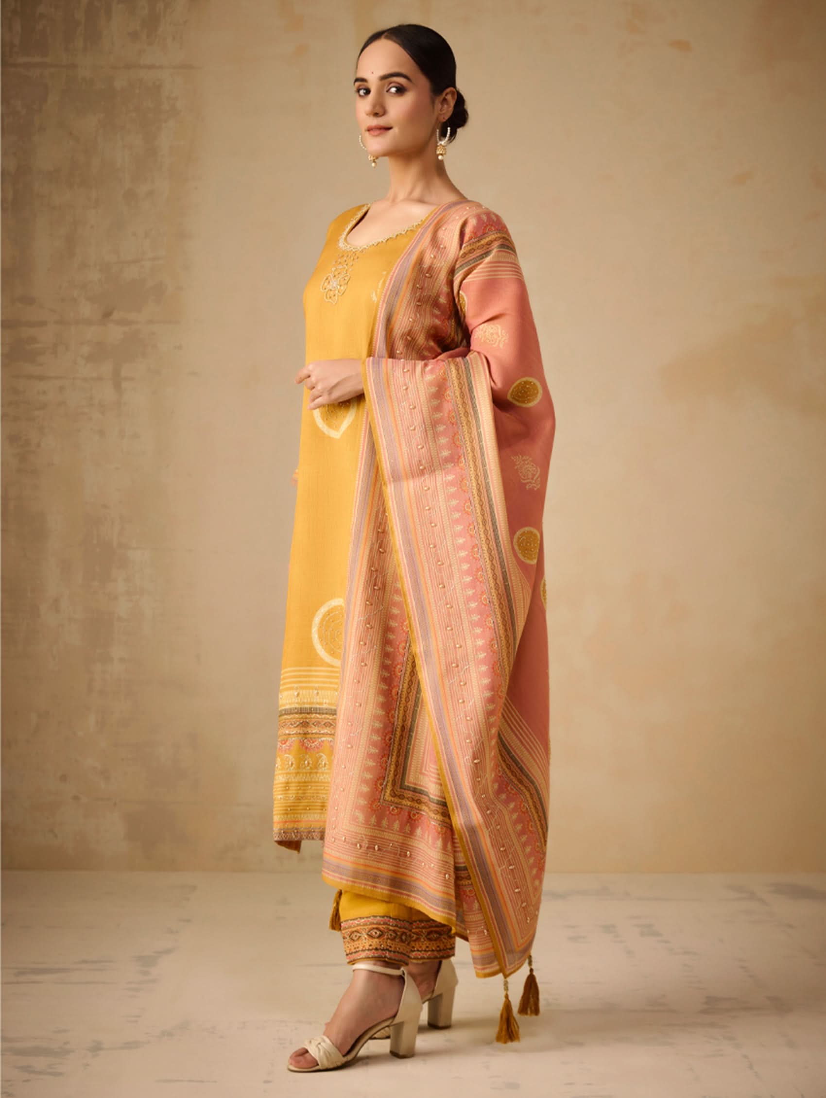 Woman wearing a yellow outfit with a pink and gold dupatta against a beige background