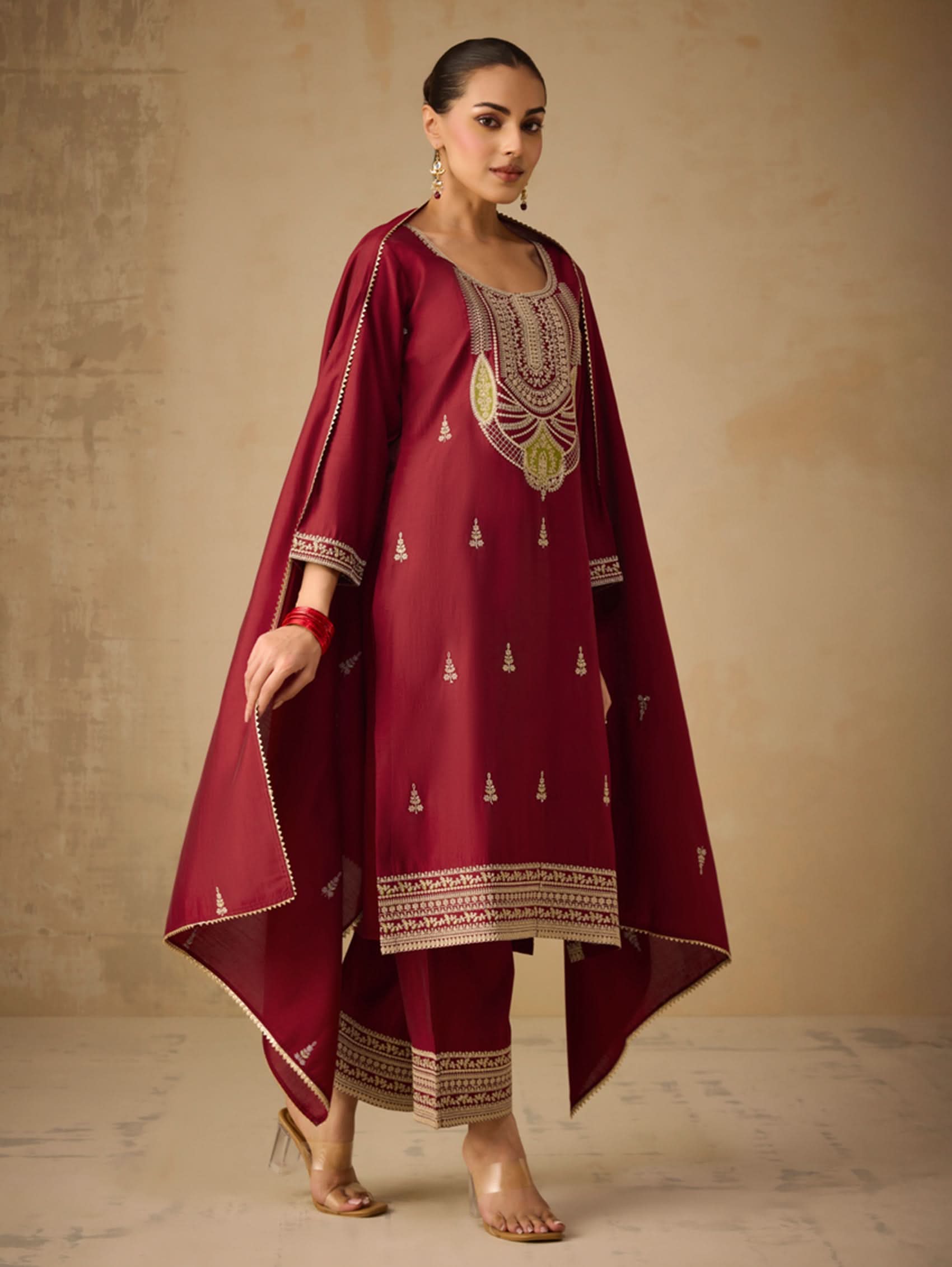Woman wearing a maroon traditional outfit with gold embroidery against a beige background