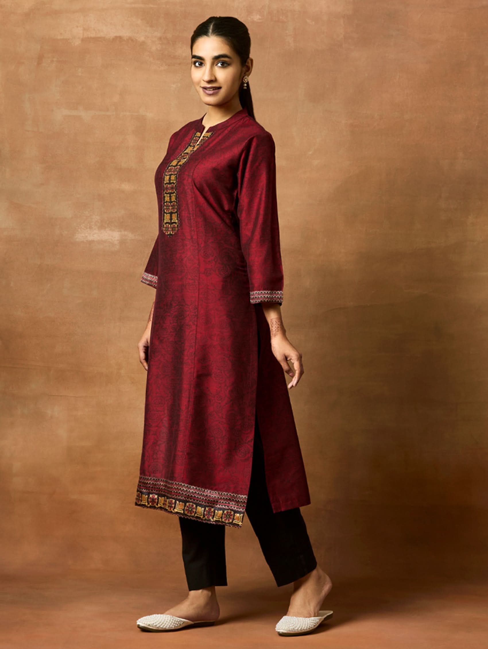 Woman wearing a maroon traditional outfit with gold embroidery against a brown background