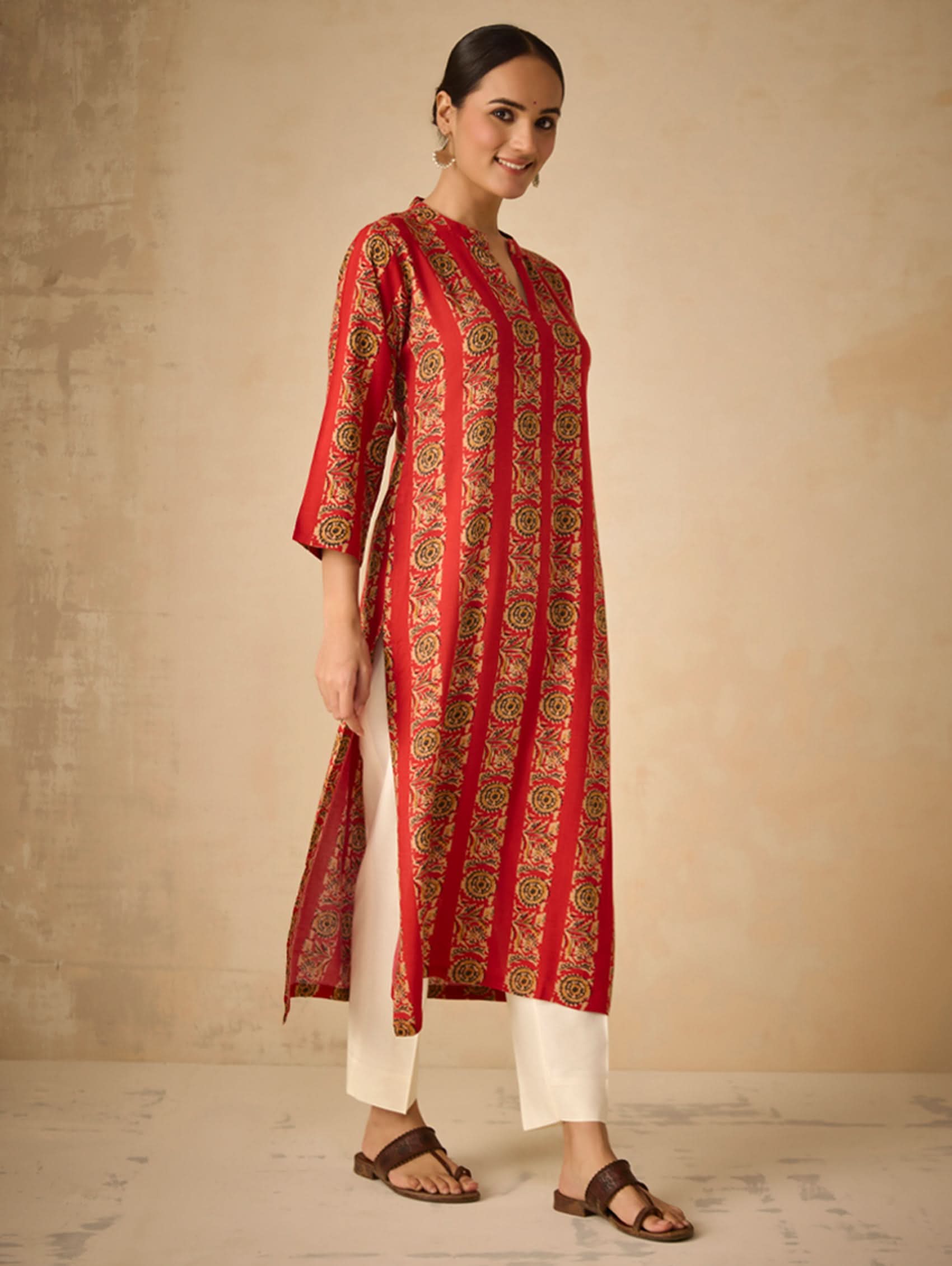 Woman wearing a red and gold patterned kurta with white pants against a beige background