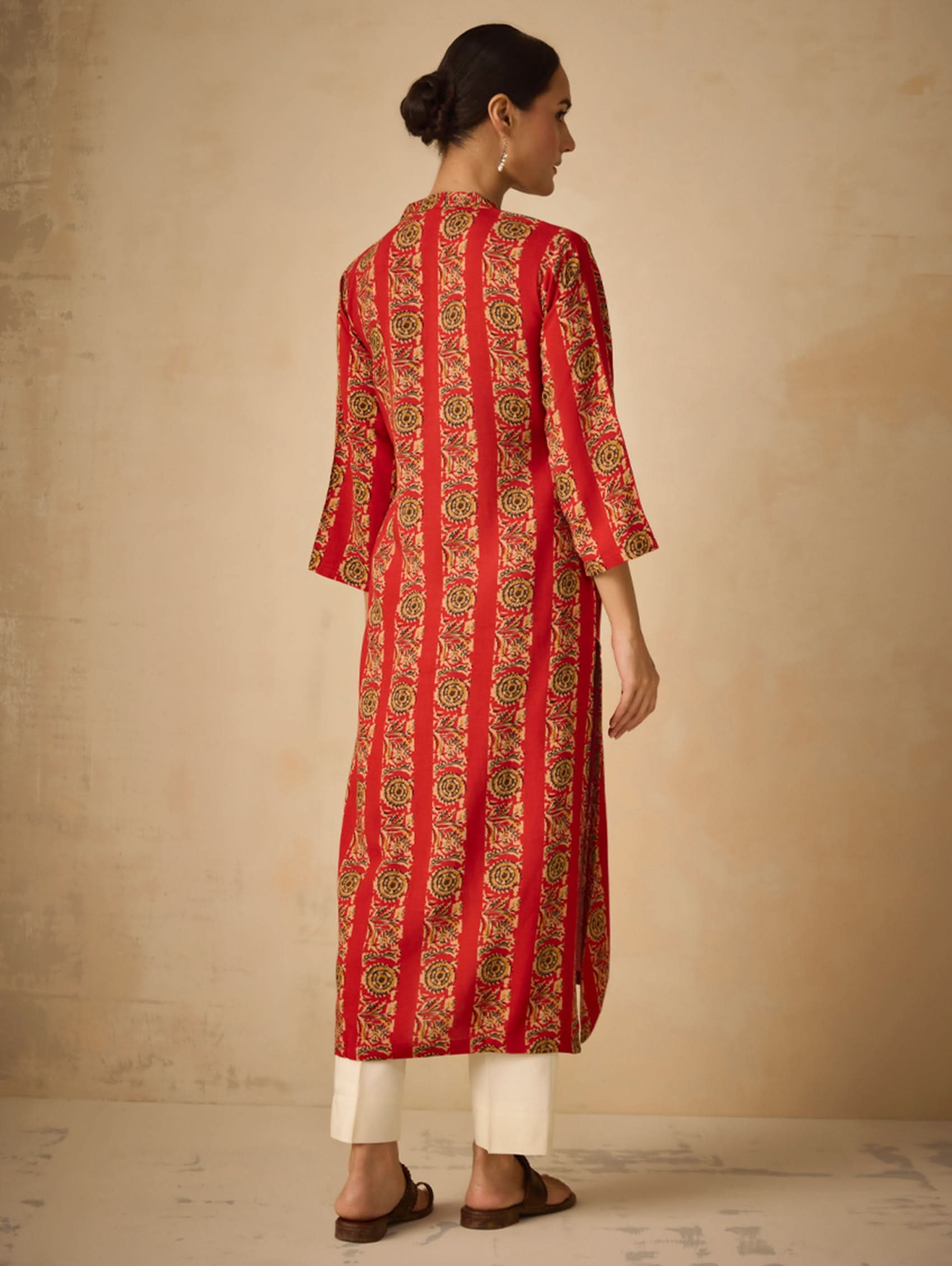Woman wearing a red and gold patterned kurta with white pants against a beige background