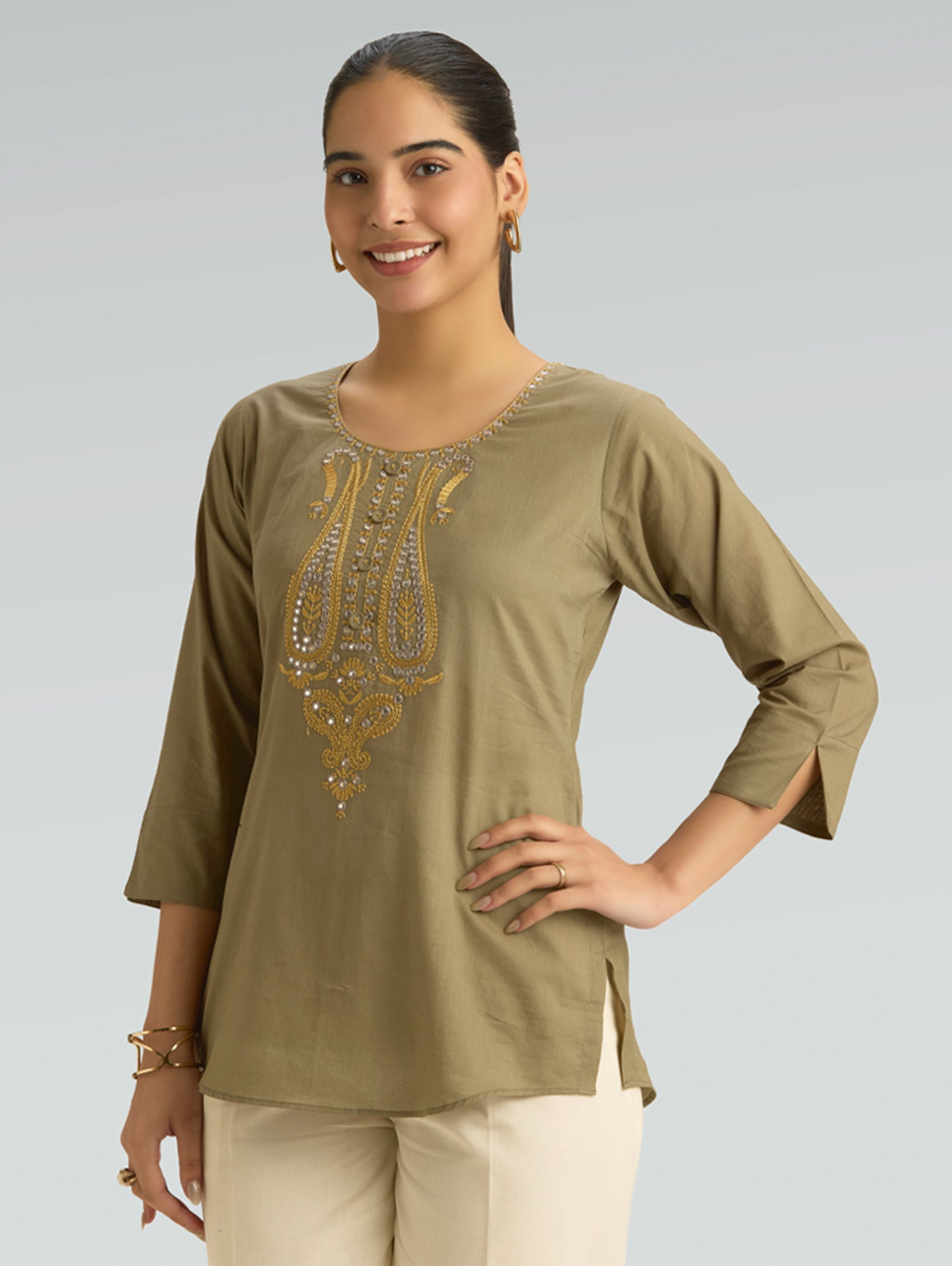 Woman wearing an olive green top with gold embroidery on a plain background