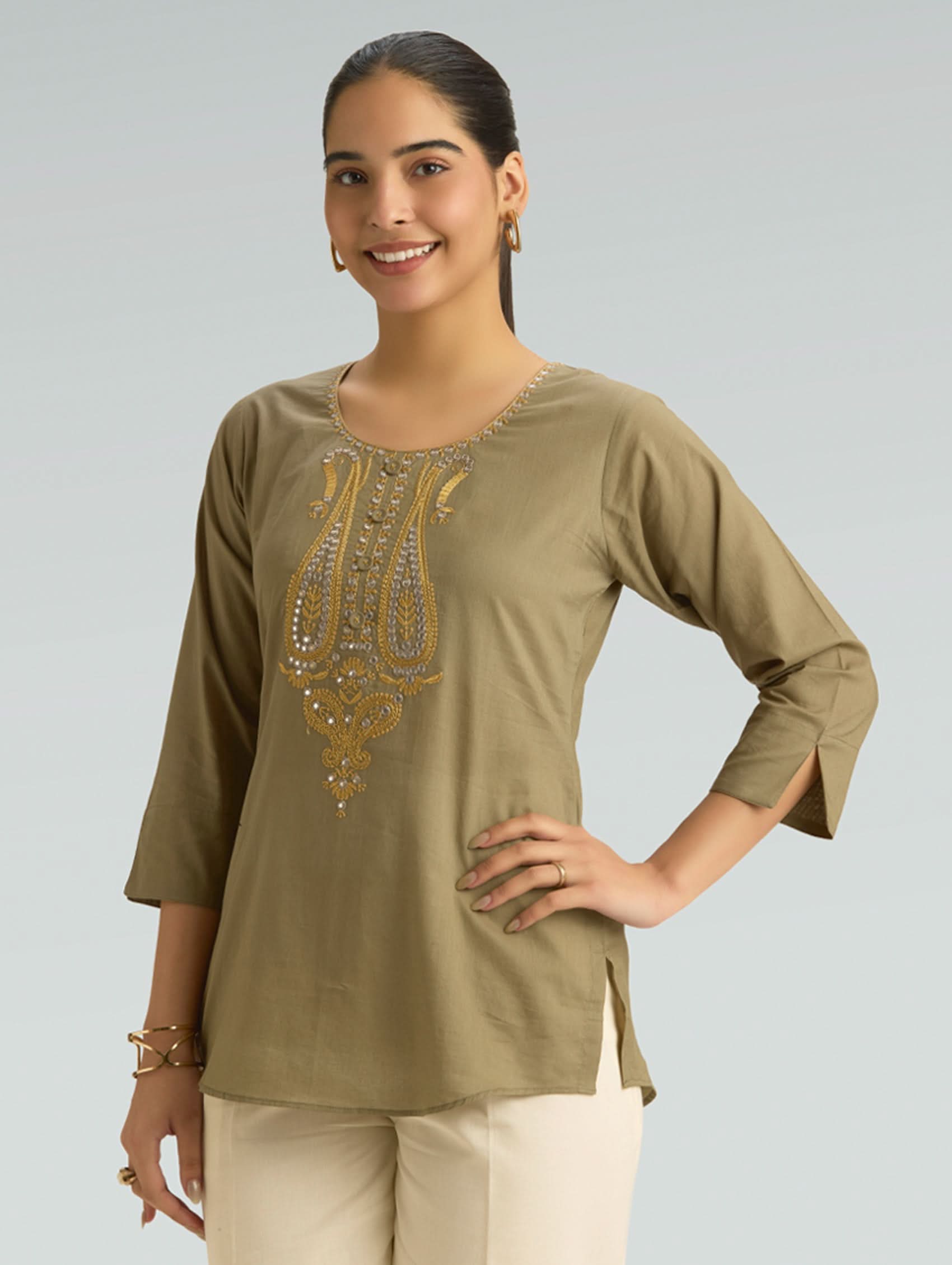 Woman wearing an olive green top with gold embroidery on a plain background