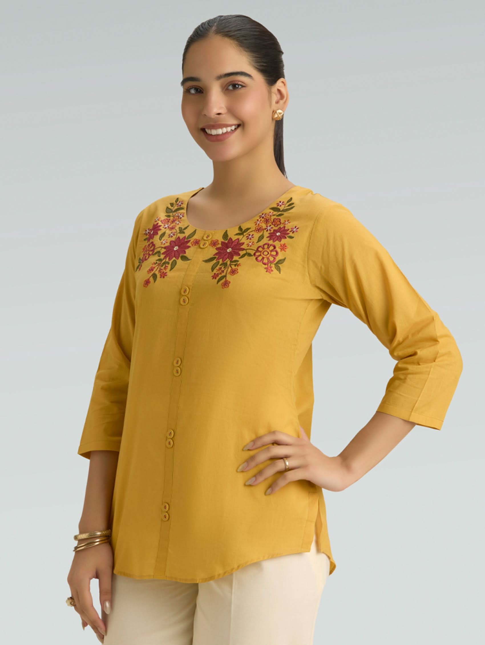 Woman wearing a mustard yellow top with floral embroidery on a gray background