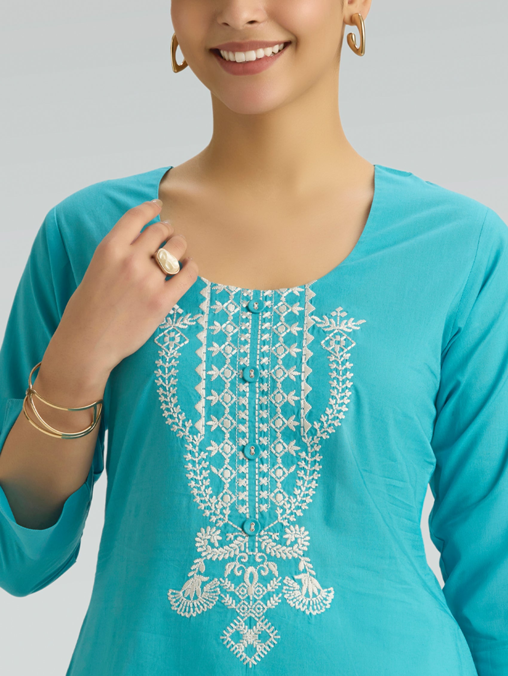 Woman wearing a turquoise top with white embroidery on a plain background