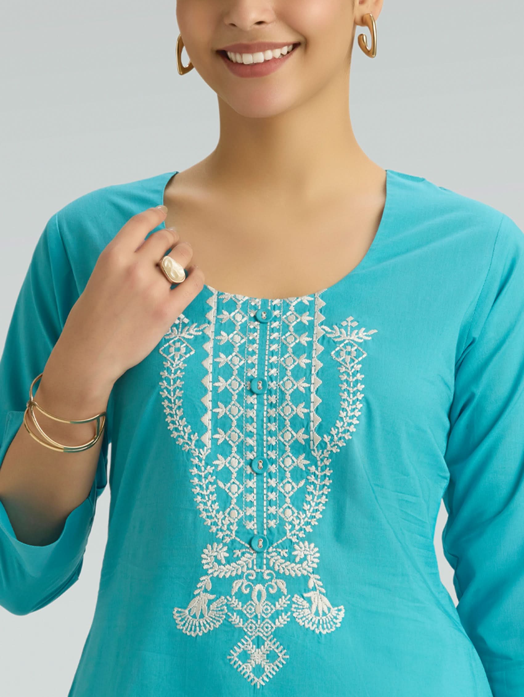 Woman wearing a turquoise top with white embroidery on a plain background