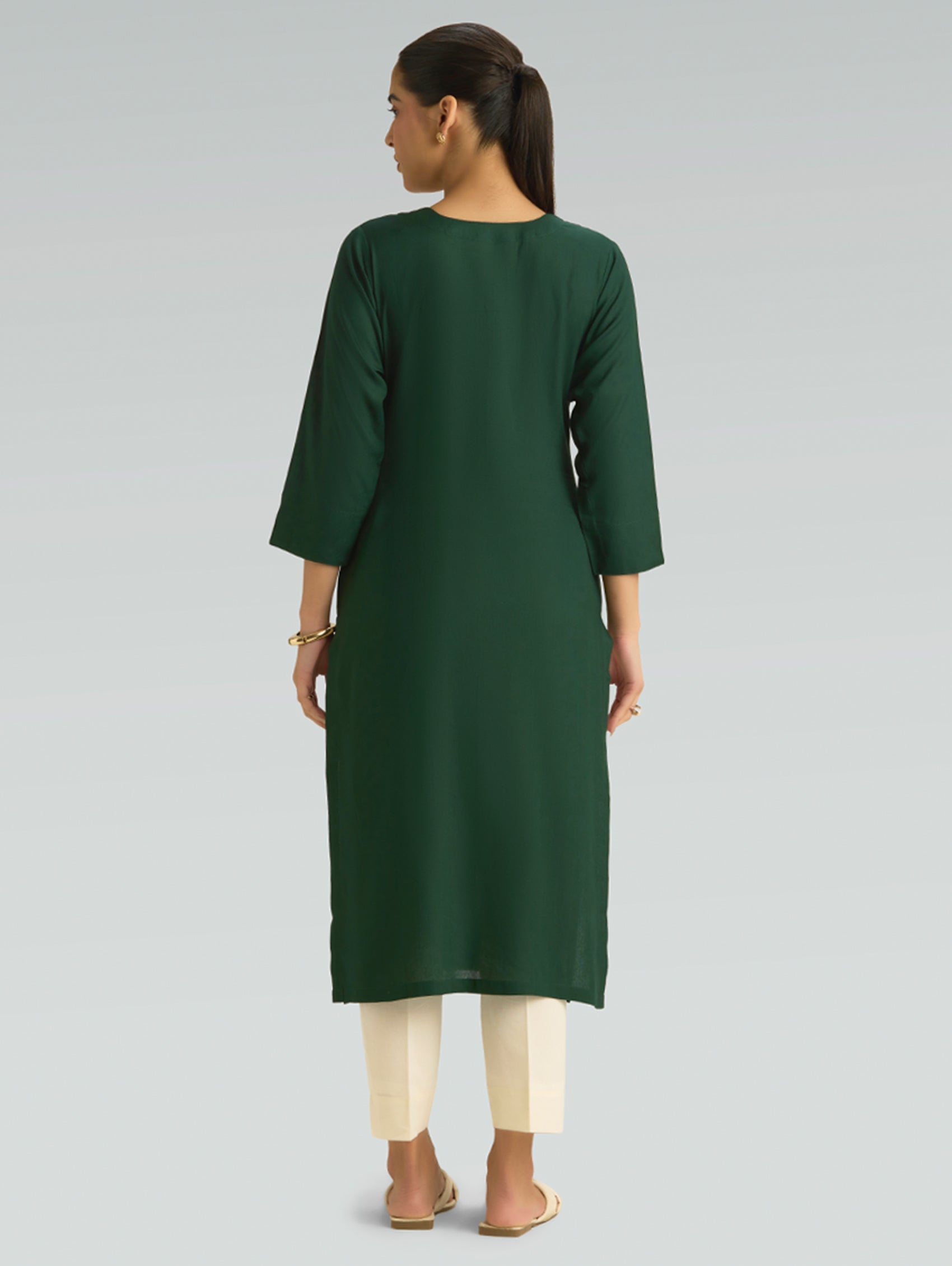 “A woman dressed in a green kurta paired with white pants, captured in a graceful back pose on a light gray background.”