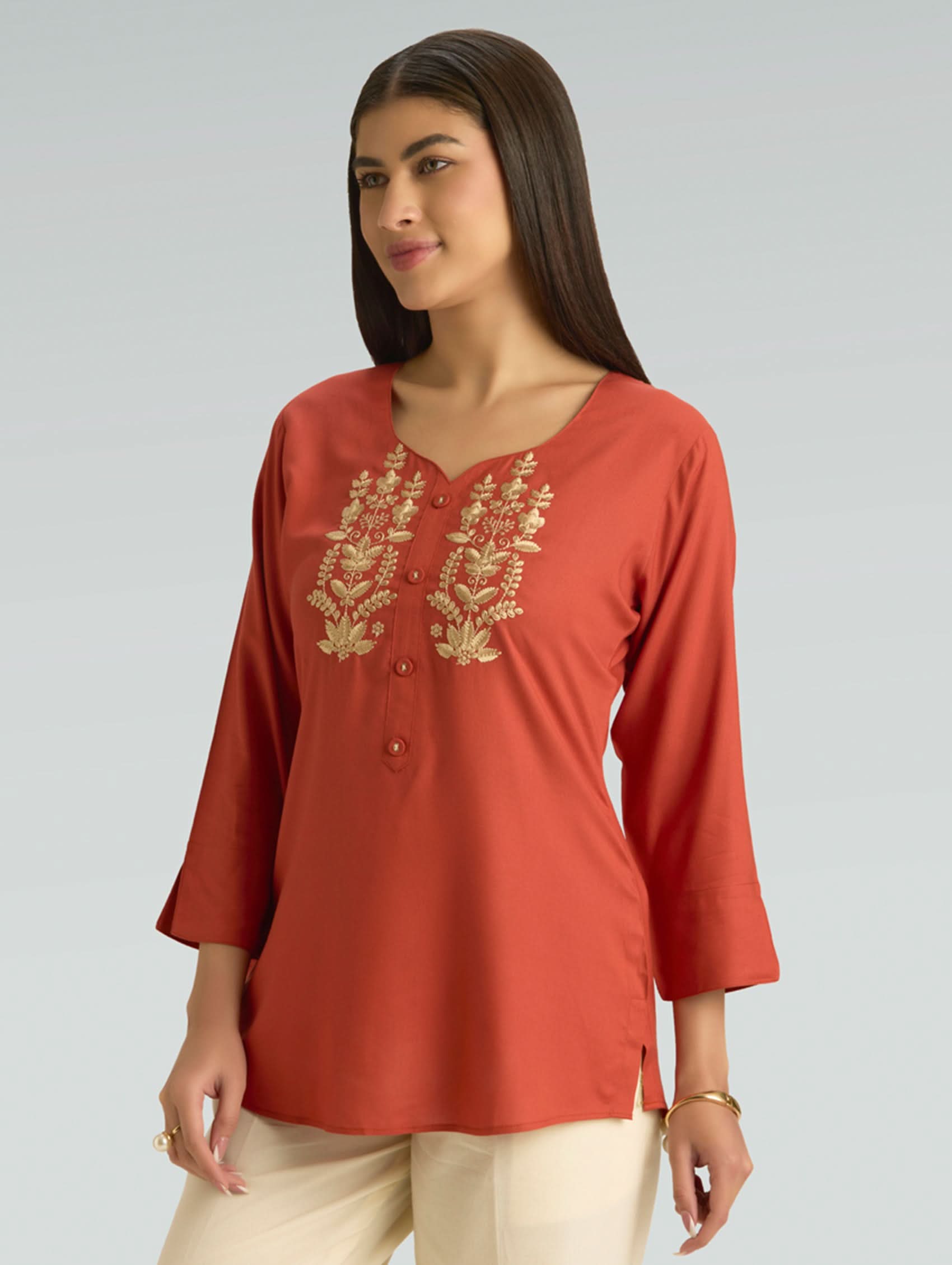 Woman wearing a rust-colored top with gold embroidery on a gray background