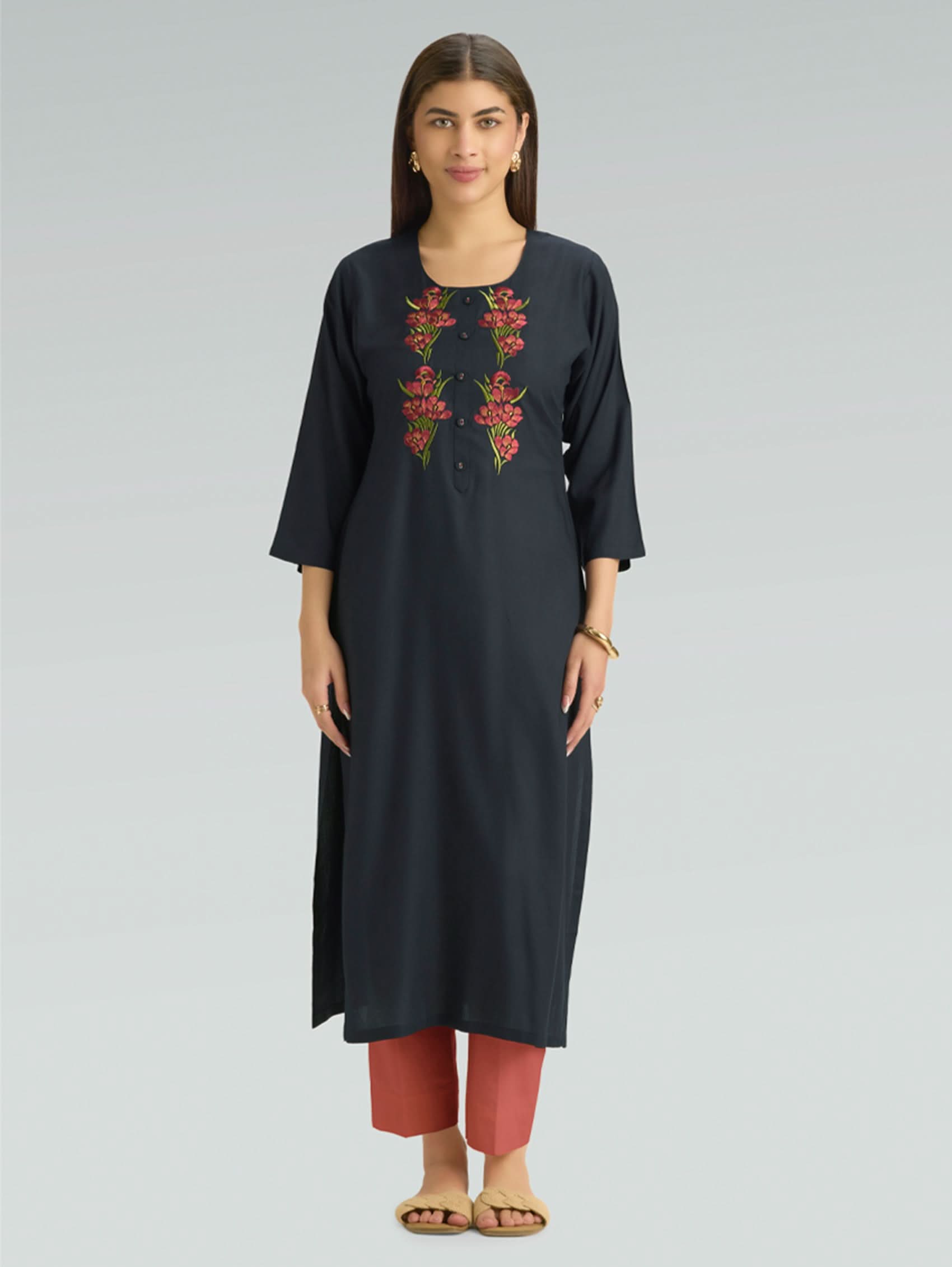 A woman wearing a straight-fit black kurta with a round neckline, adorned with floral embroidery, set against a soft gray background.