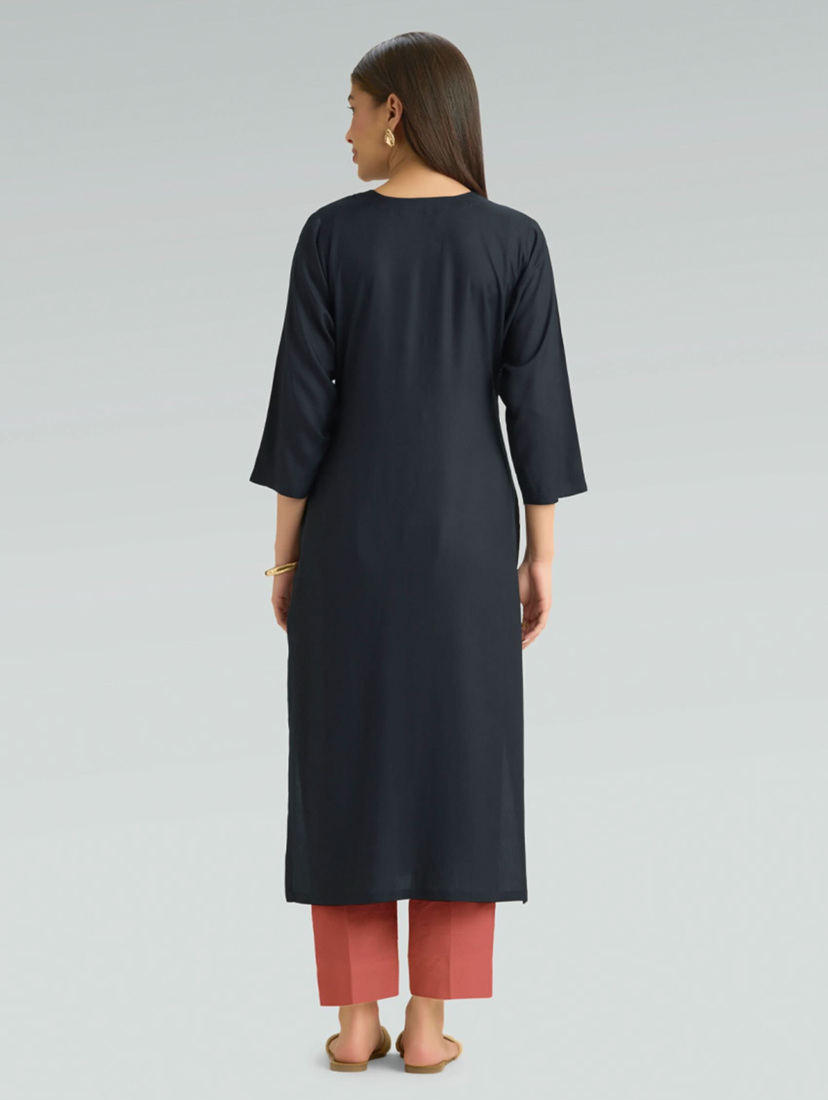 A woman in a straight-fit black kurta with a round neckline and floral embroidery, shown in a back pose against a soft gray background.