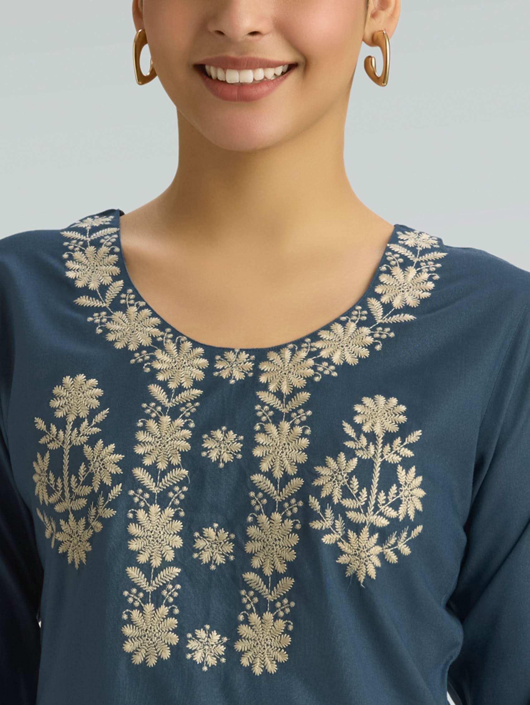 Woman wearing a blue top with gold floral embroidery on a gray background