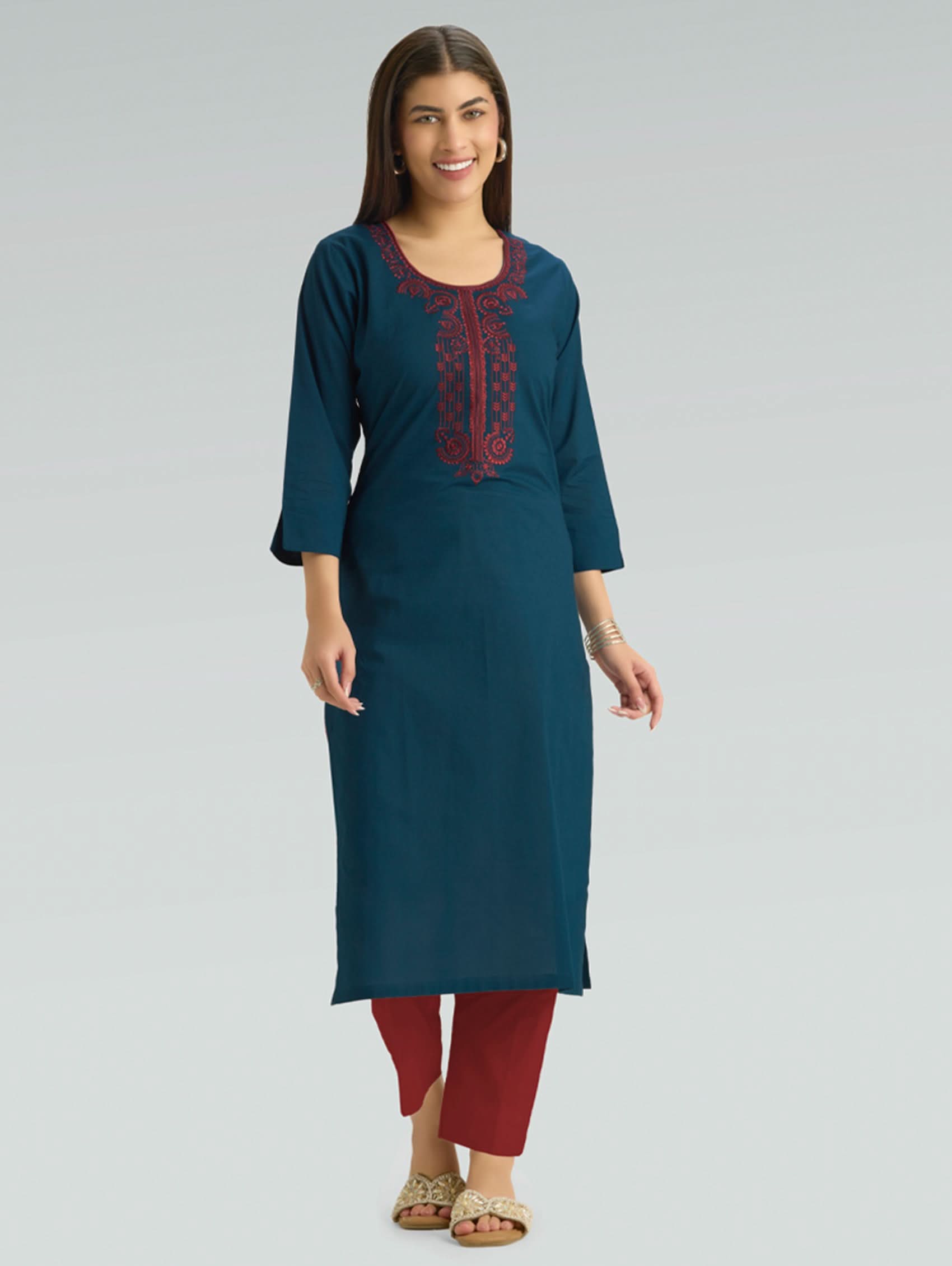 A woman wearing a straight-fit teal kurta with a round neckline, adorned with intricate red embroidery, set against a soft gray background.