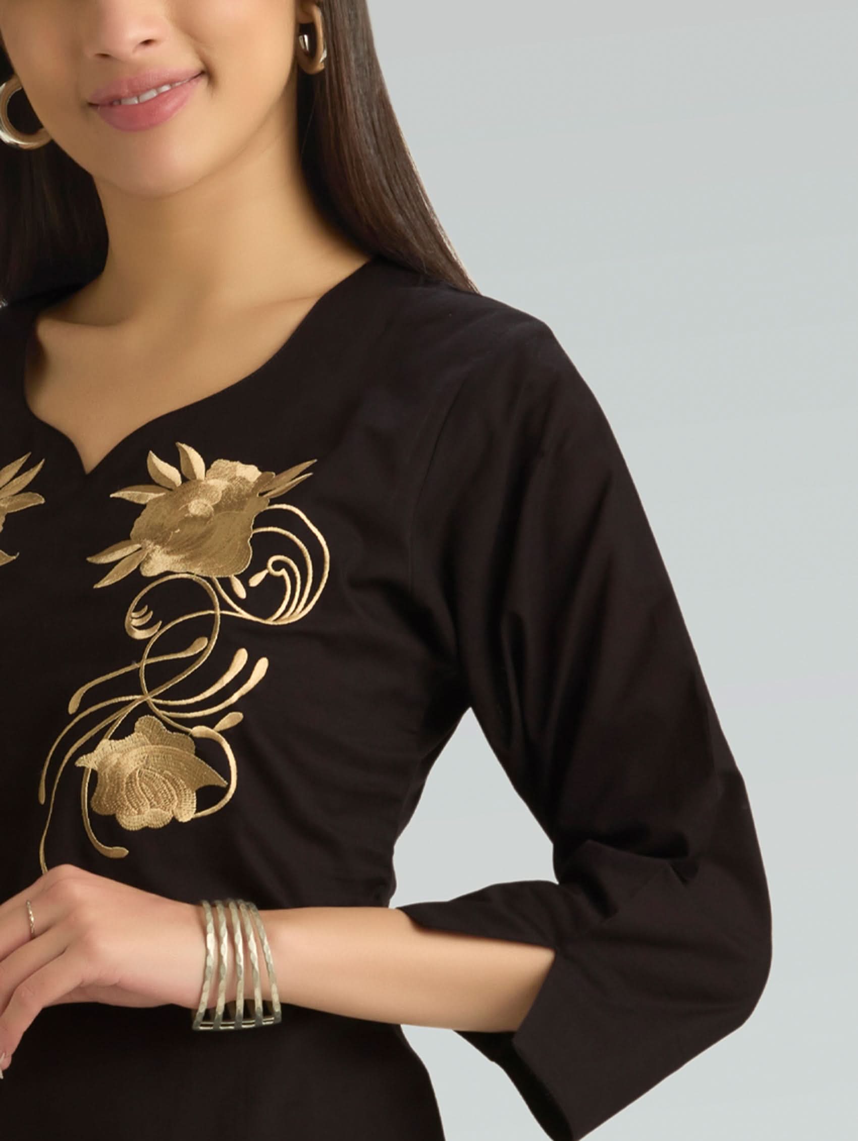 Woman wearing a black top with gold floral embroidery on a plain background