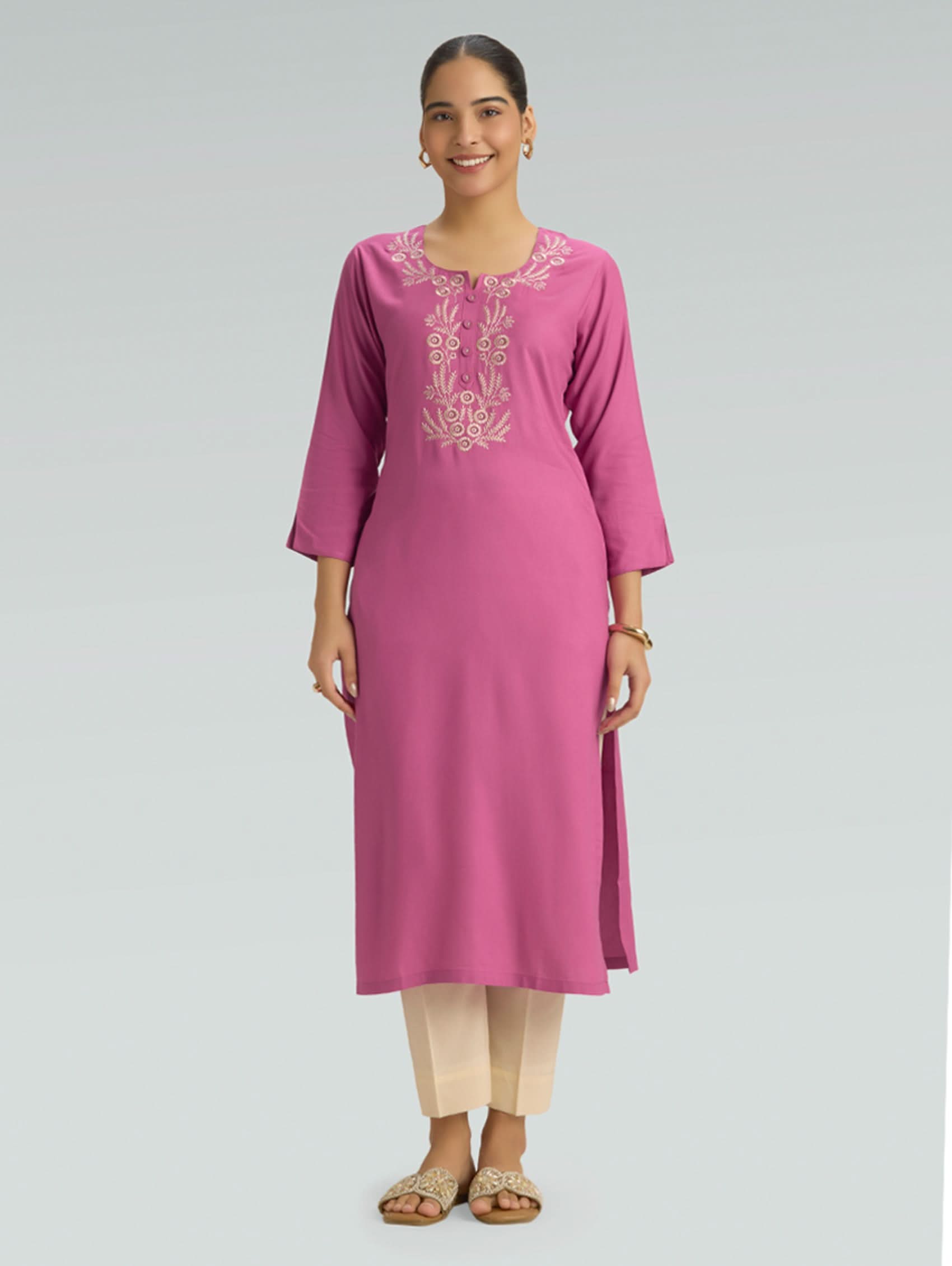 Woman wearing a pink embroidered kurta with a round neckline and 3/4 sleeves, paired with beige pants on a gray background.