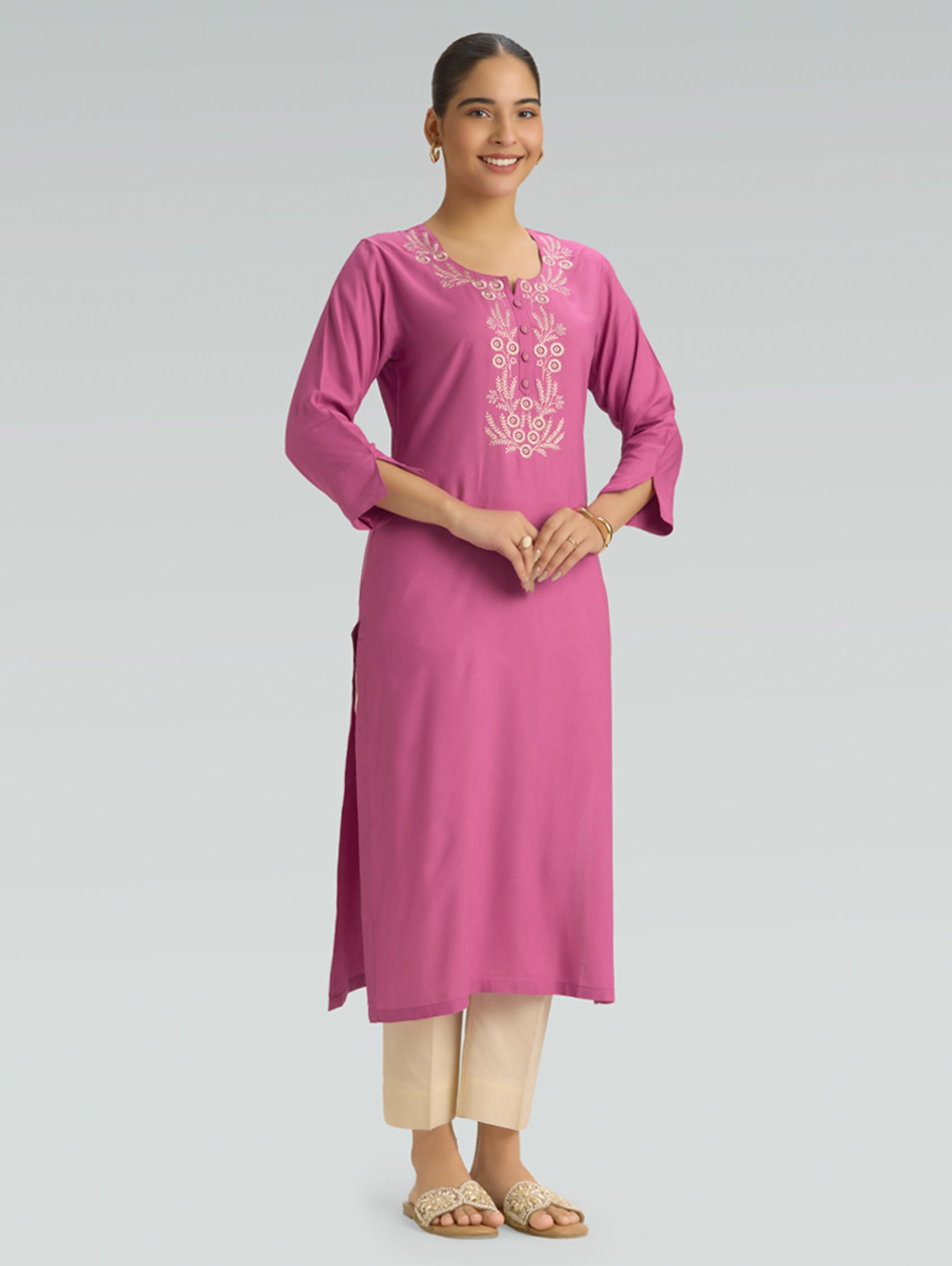 A woman is wearing a pink kurta with white embroidery and a side slit, along with 3/4 sleeves, against a grey background.