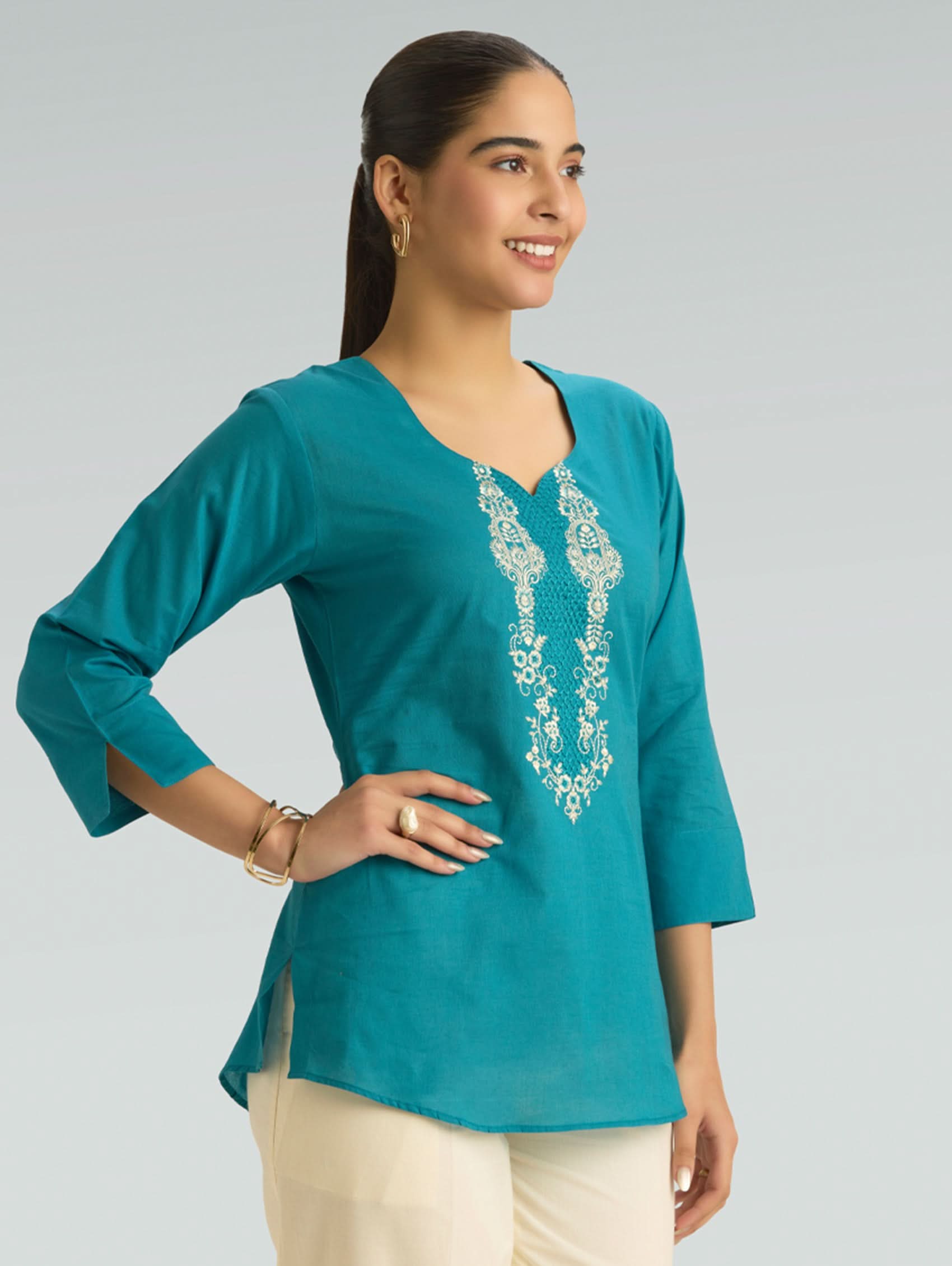 Woman wearing a teal top with floral embroidery on a gray background