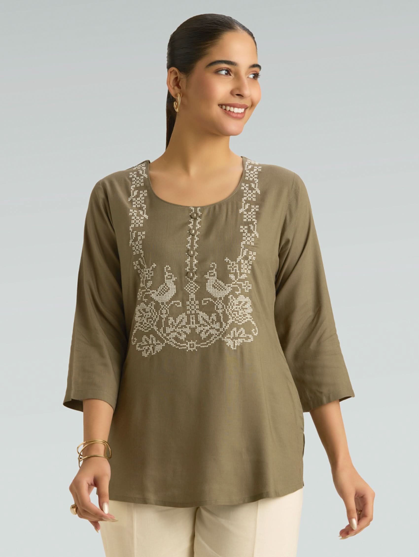 Woman wearing an olive green top with embroidery on a plain background