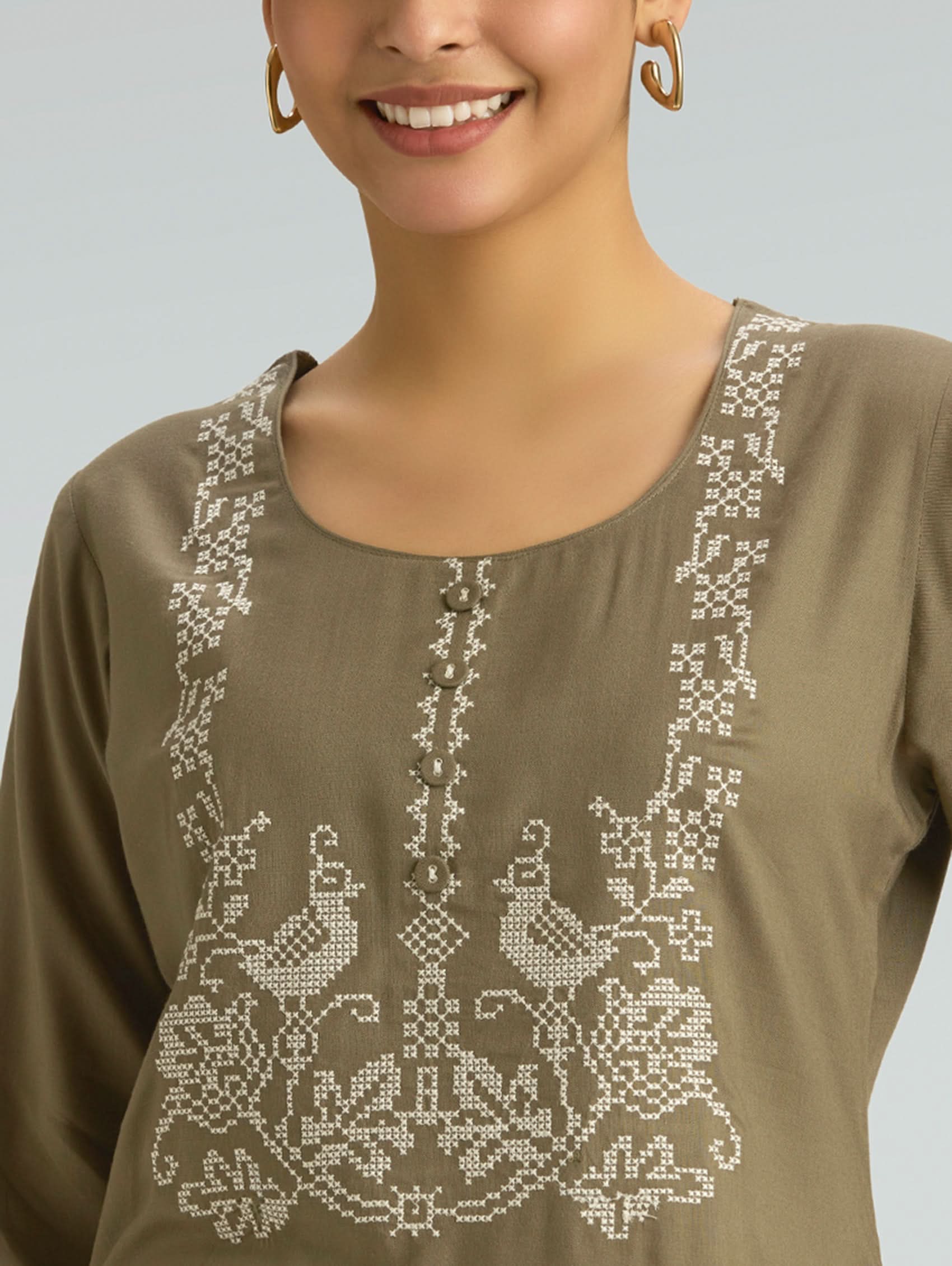 Woman wearing a brown top with intricate white embroidery on a plain background