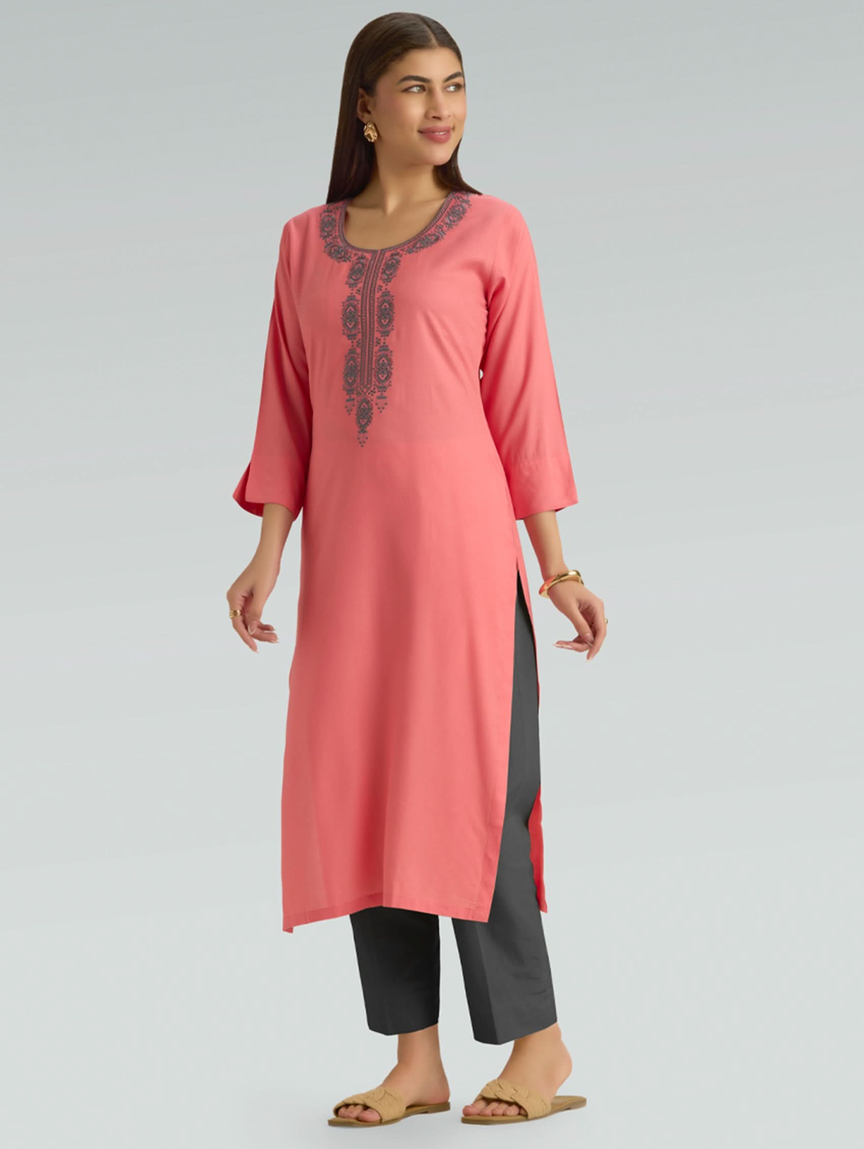 A woman wearing a flamingo pink kurta with grey embroidery on the kurta and black pants on a grey background