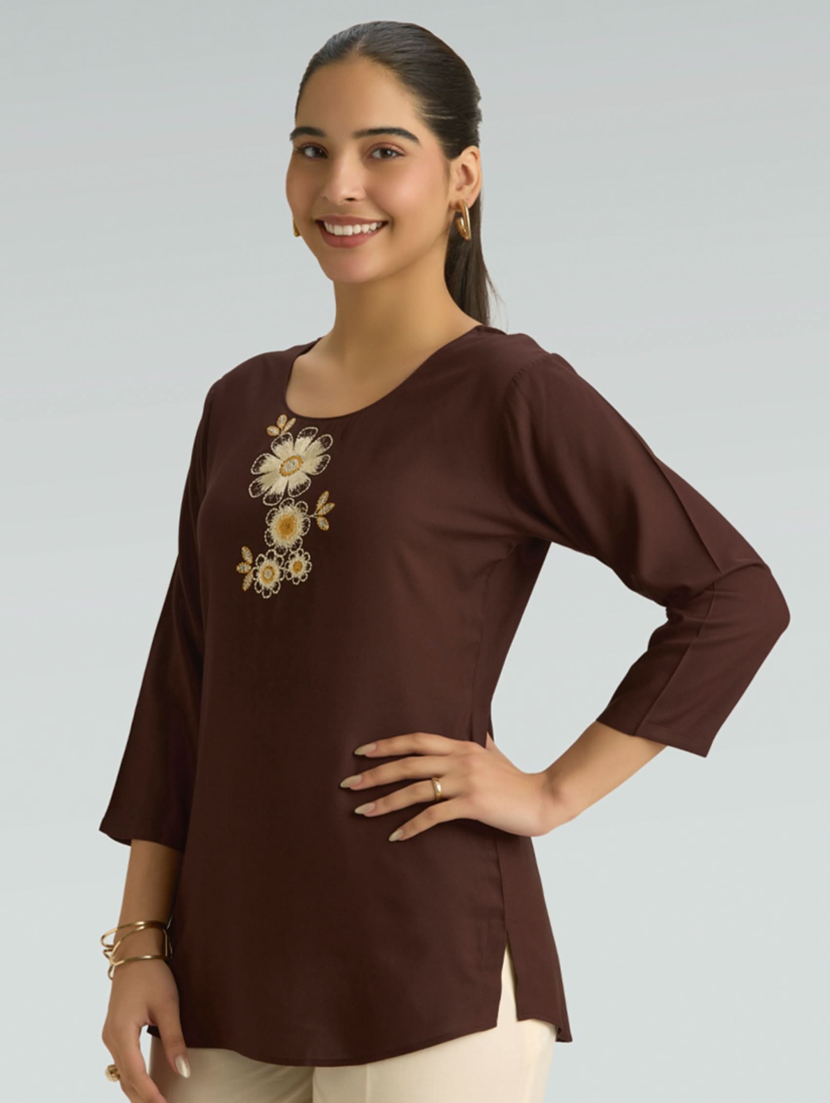 Woman wearing a brown long-sleeve top with floral embroidery on a gray background