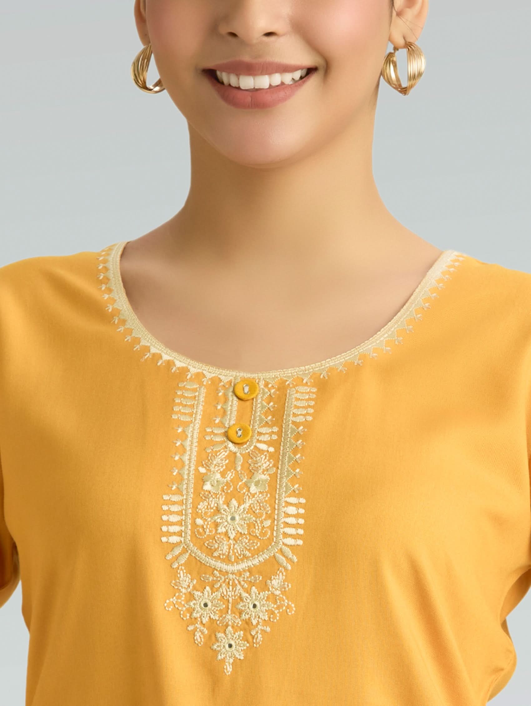 Woman wearing a mustard yellow top with intricate white embroidery on a gray background