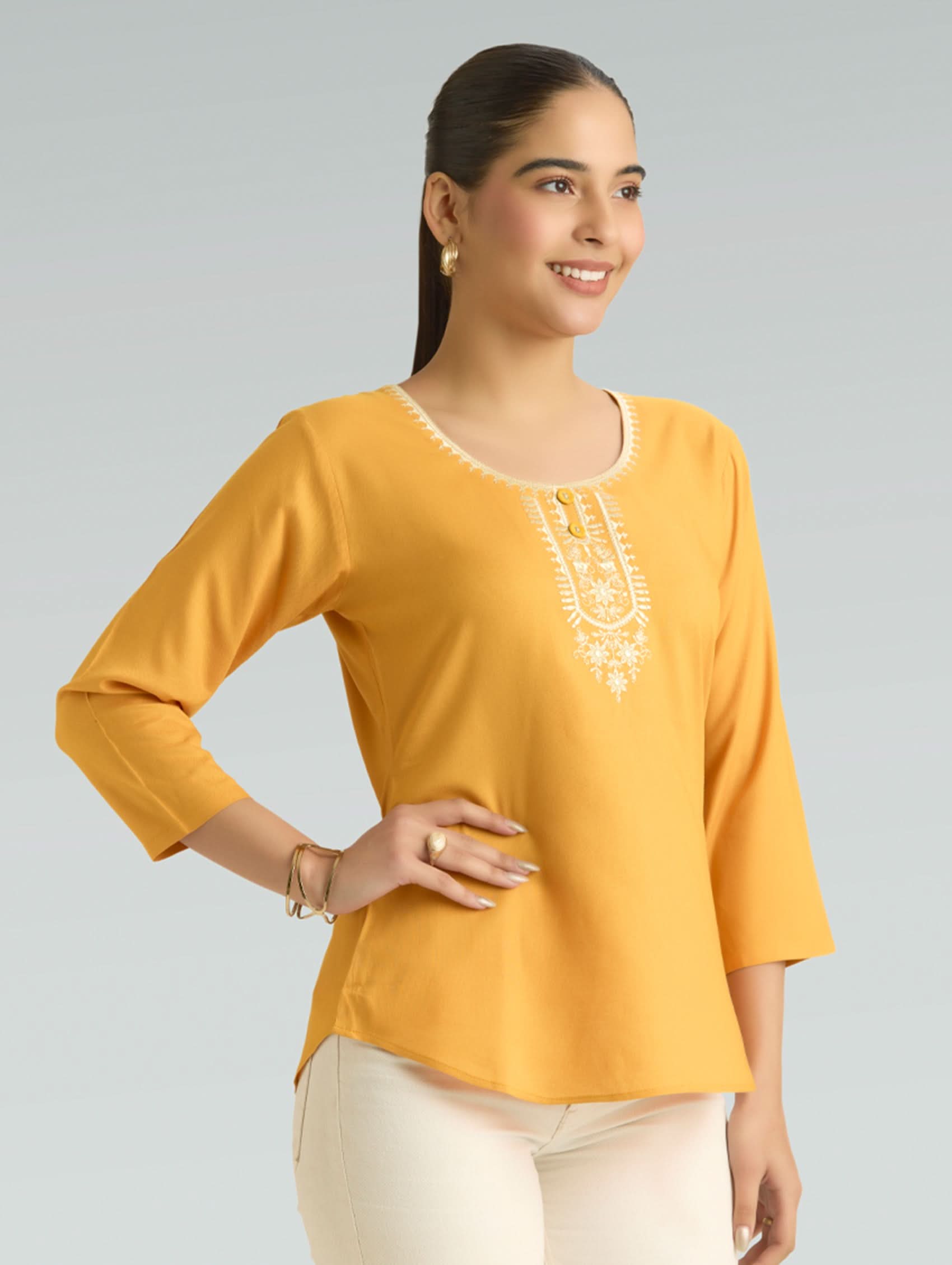 Woman wearing a mustard yellow top with white embroidery on a gray background