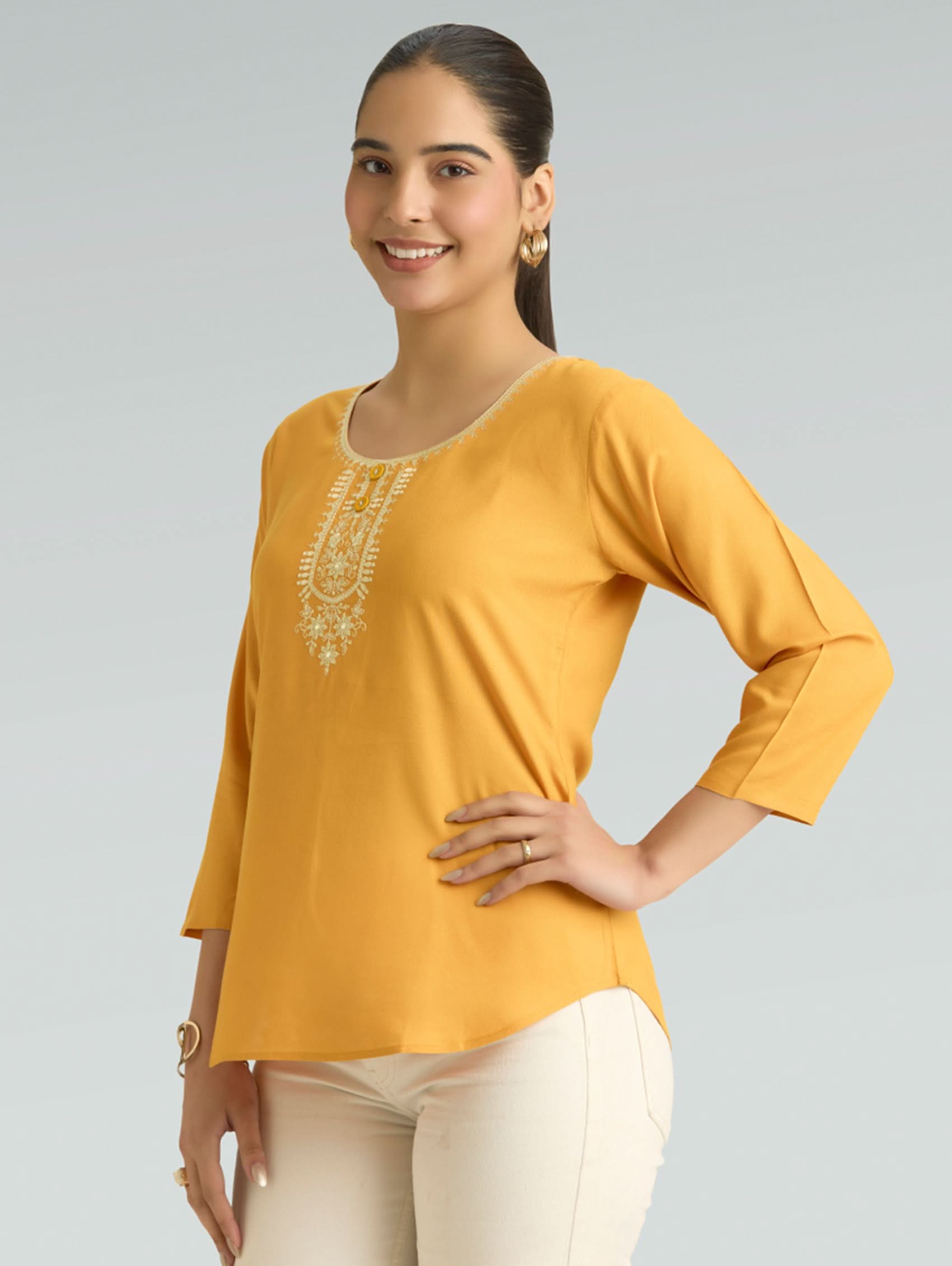 Woman wearing a mustard yellow top with white embroidery on a gray background