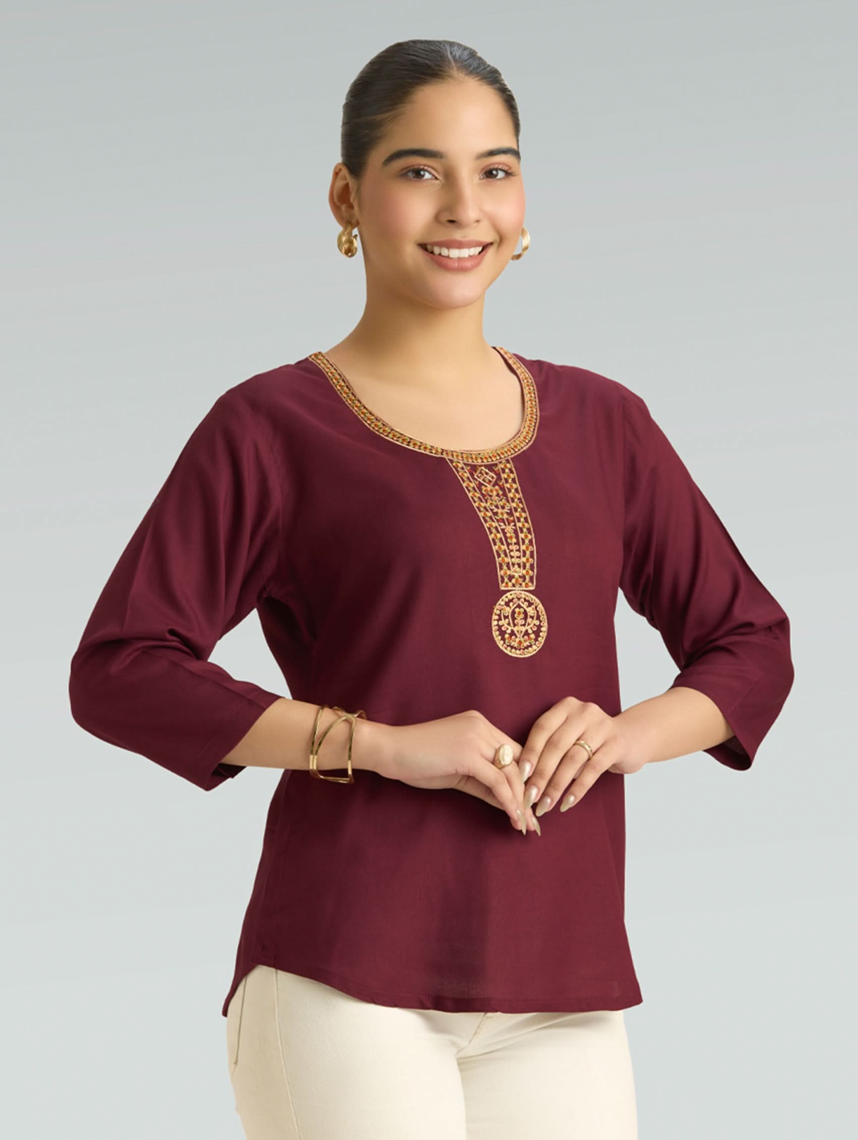 Woman wearing a maroon top with gold embroidery on a gray background
