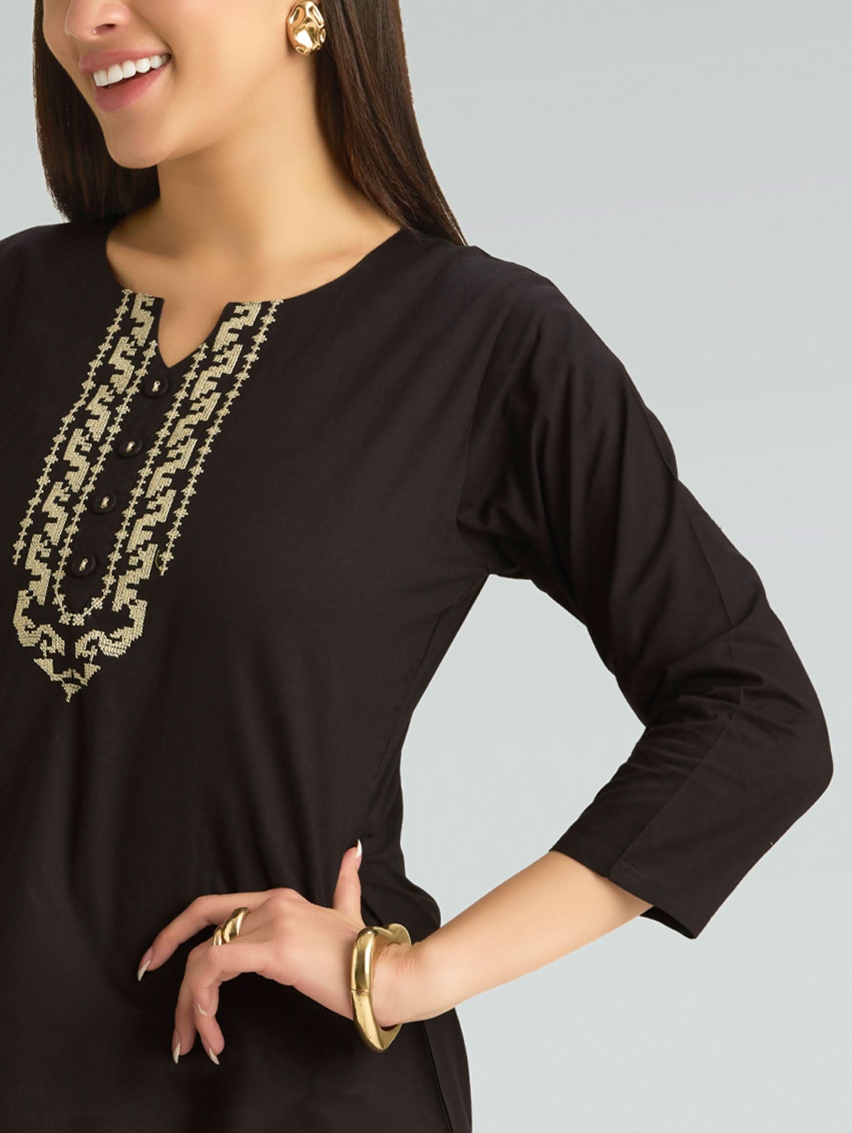Woman wearing a black long-sleeve top with gold embroidery on a gray background